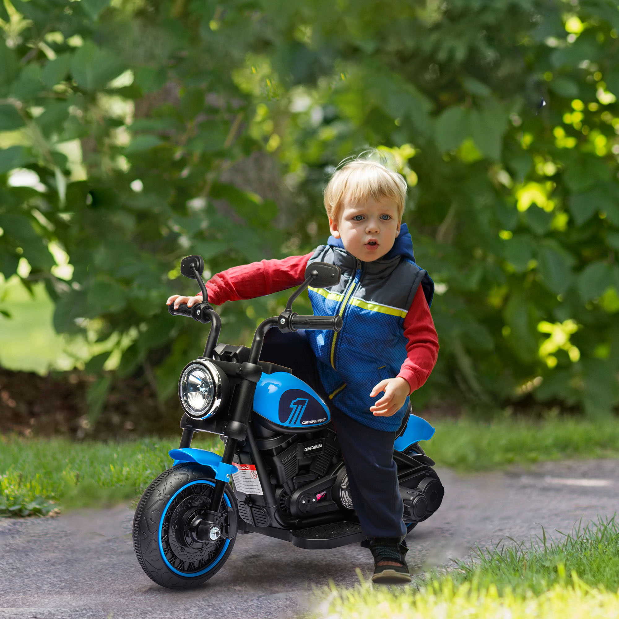 Un bambino piccolo su una moto giocattolo blu, che guarda la fotocamera. Lo sfondo è un'area verde e erbosa.