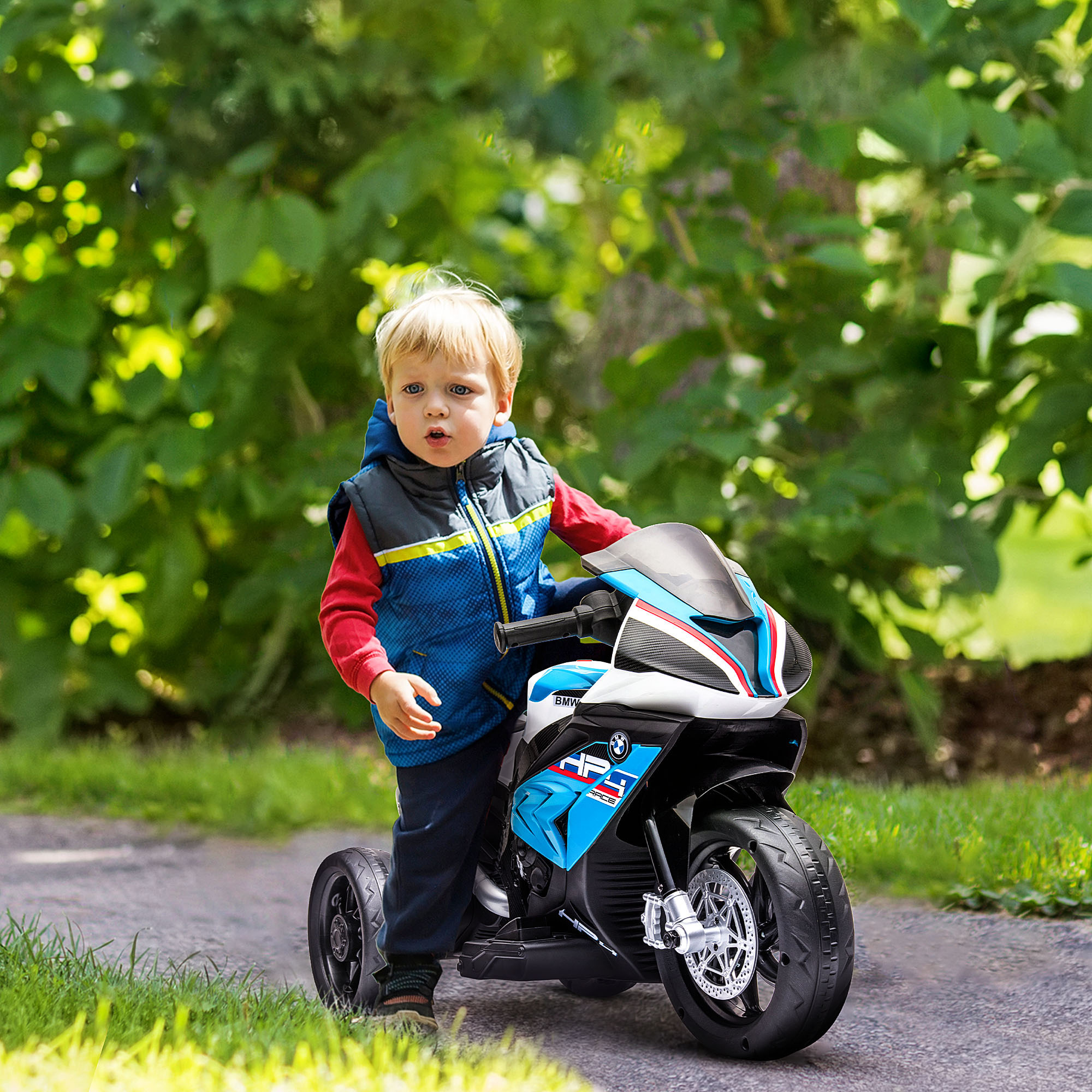 Un ragazzino è in piedi accanto a una moto giocattolo blu e bianca su un sentiero in un giardino verde.