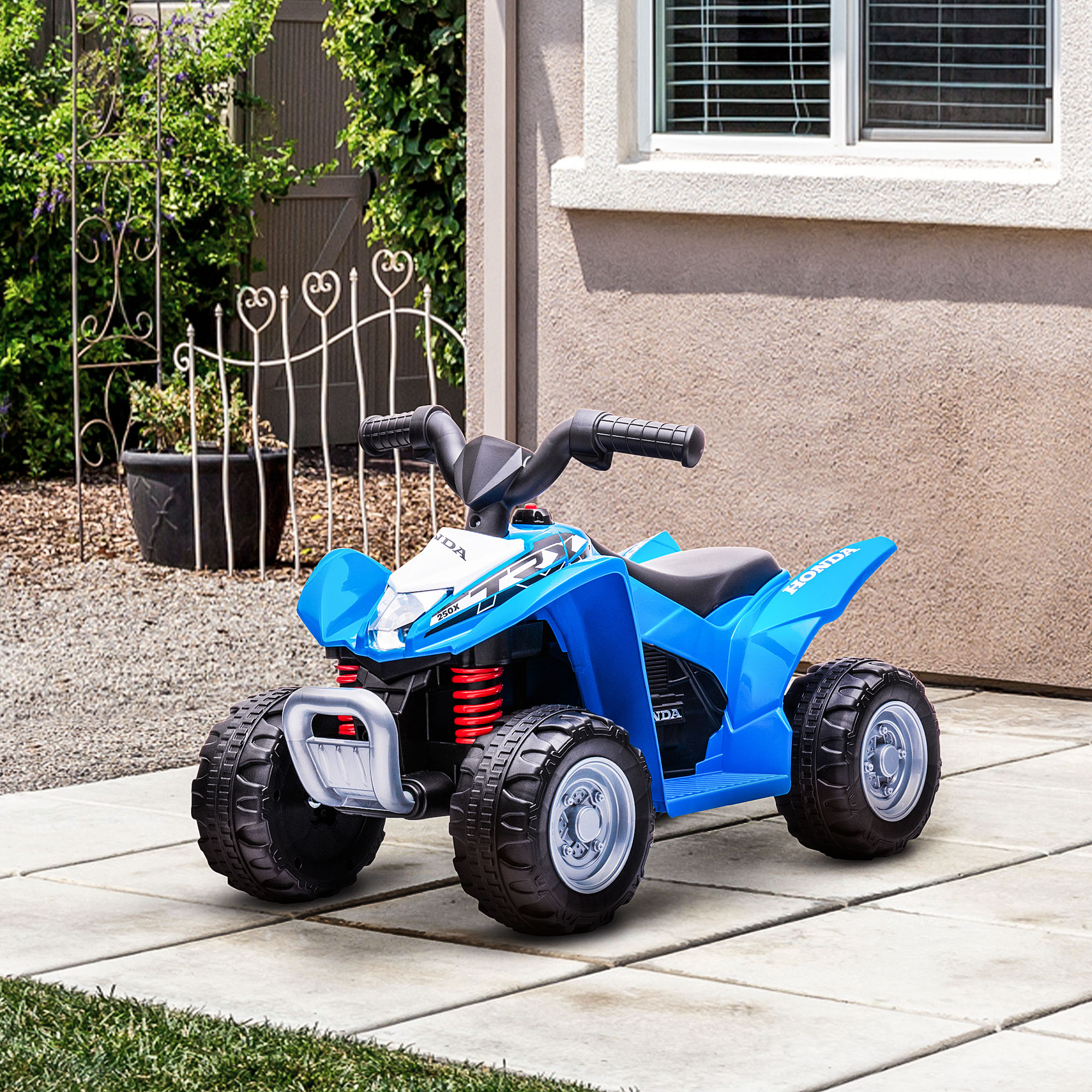 ATV giocattolo blu su una superficie pavimentata. C'è erba verde e alcuni cespugli sullo sfondo.