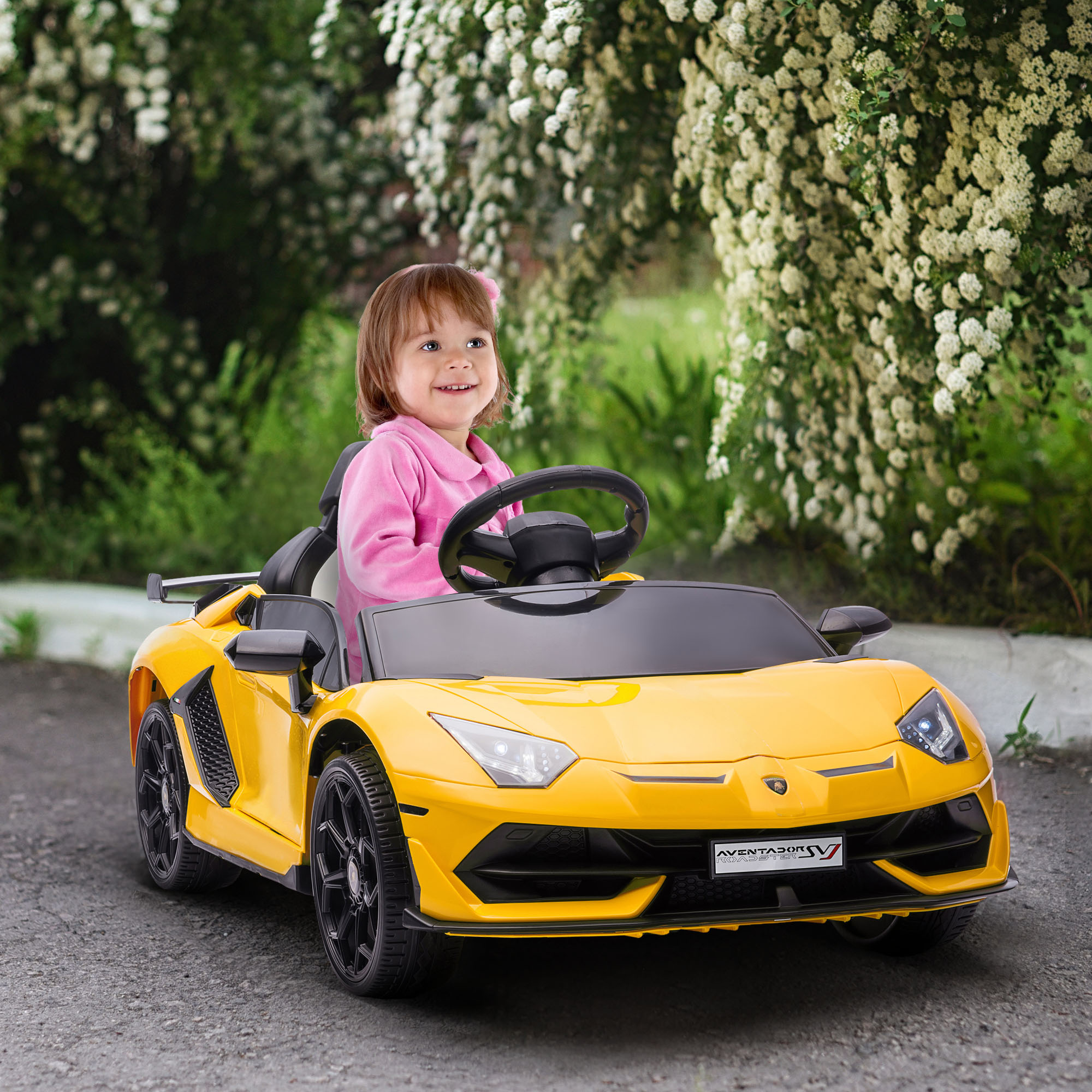 Una bambina sorride mentre guida un'auto giocattolo gialla in un giardino. Fiori bianchi sullo sfondo.