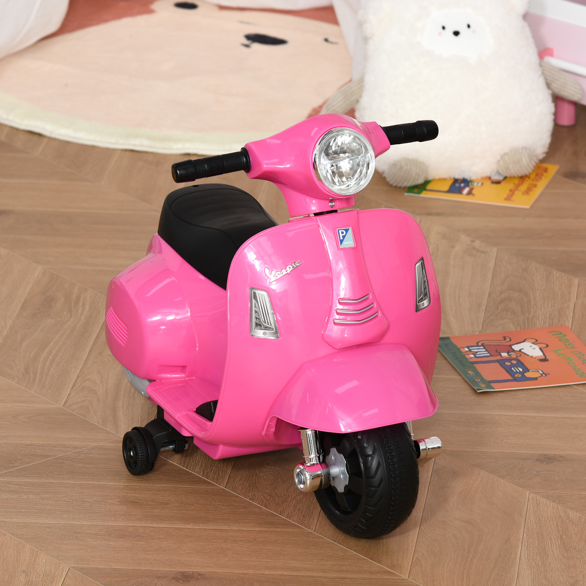 Scooter Vespa giocattolo rosa su pavimento in legno. Un peluche a forma di pecora e un libro per bambini sullo sfondo.