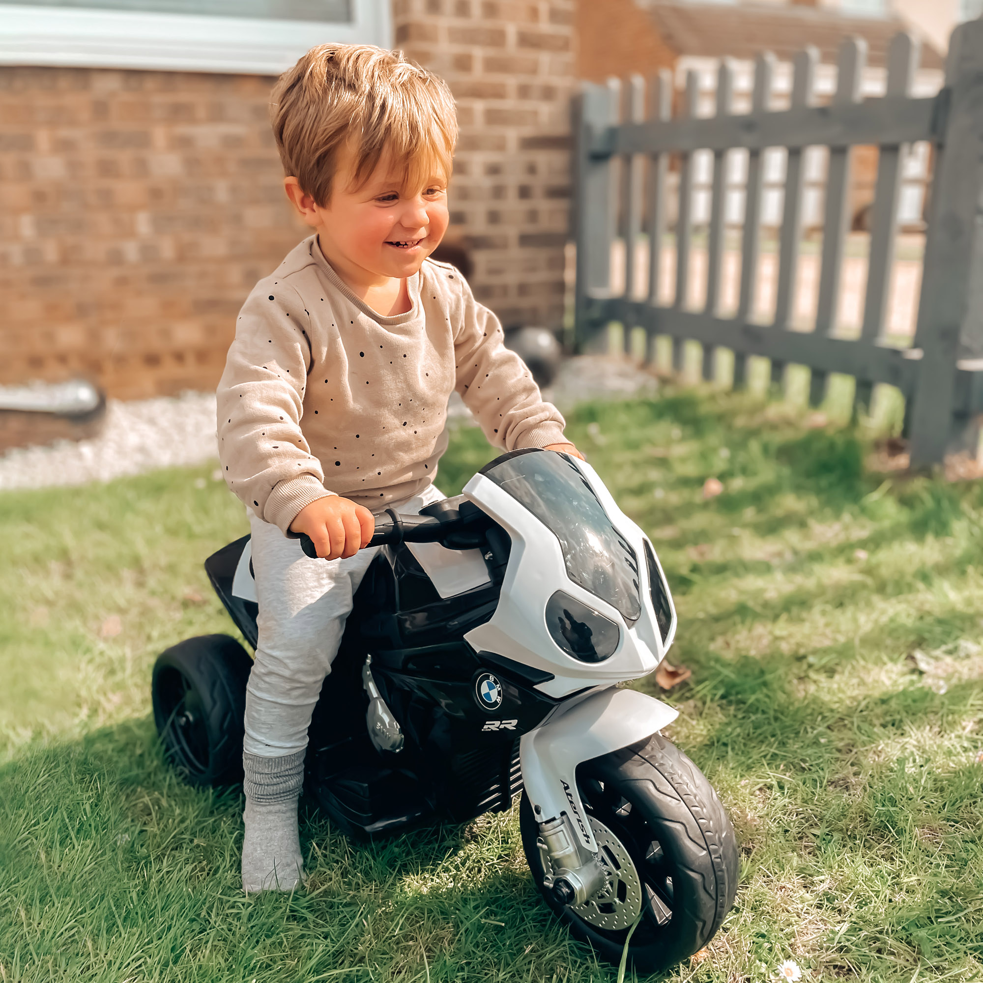 Un bambino su una moto giocattolo bianca e nera. Sta sorridendo ed è seduto sull'erba di fronte a una casa.