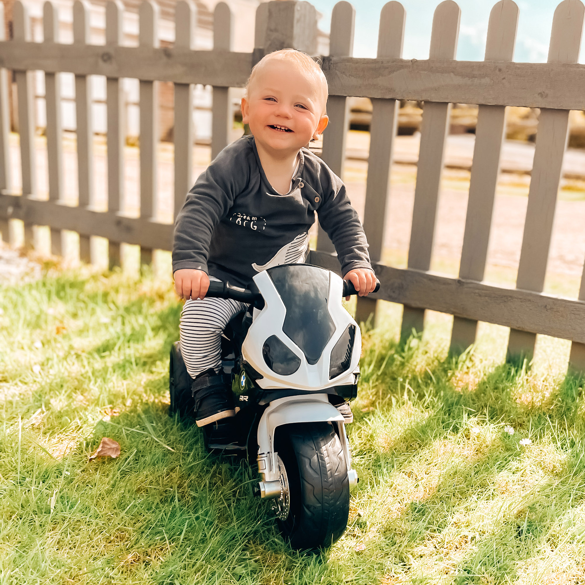Un bambino su una moto giocattolo bianca e nera, sorridente. Lo sfondo è un'area erbosa con una recinzione.