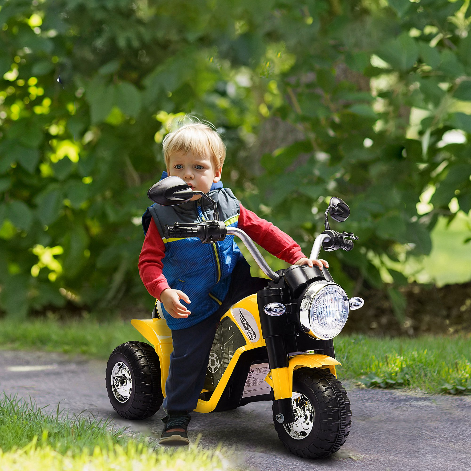 Un bambino è seduto su una moto giocattolo gialla e nera. Uno sfondo verde di fogliame è dietro di lui.