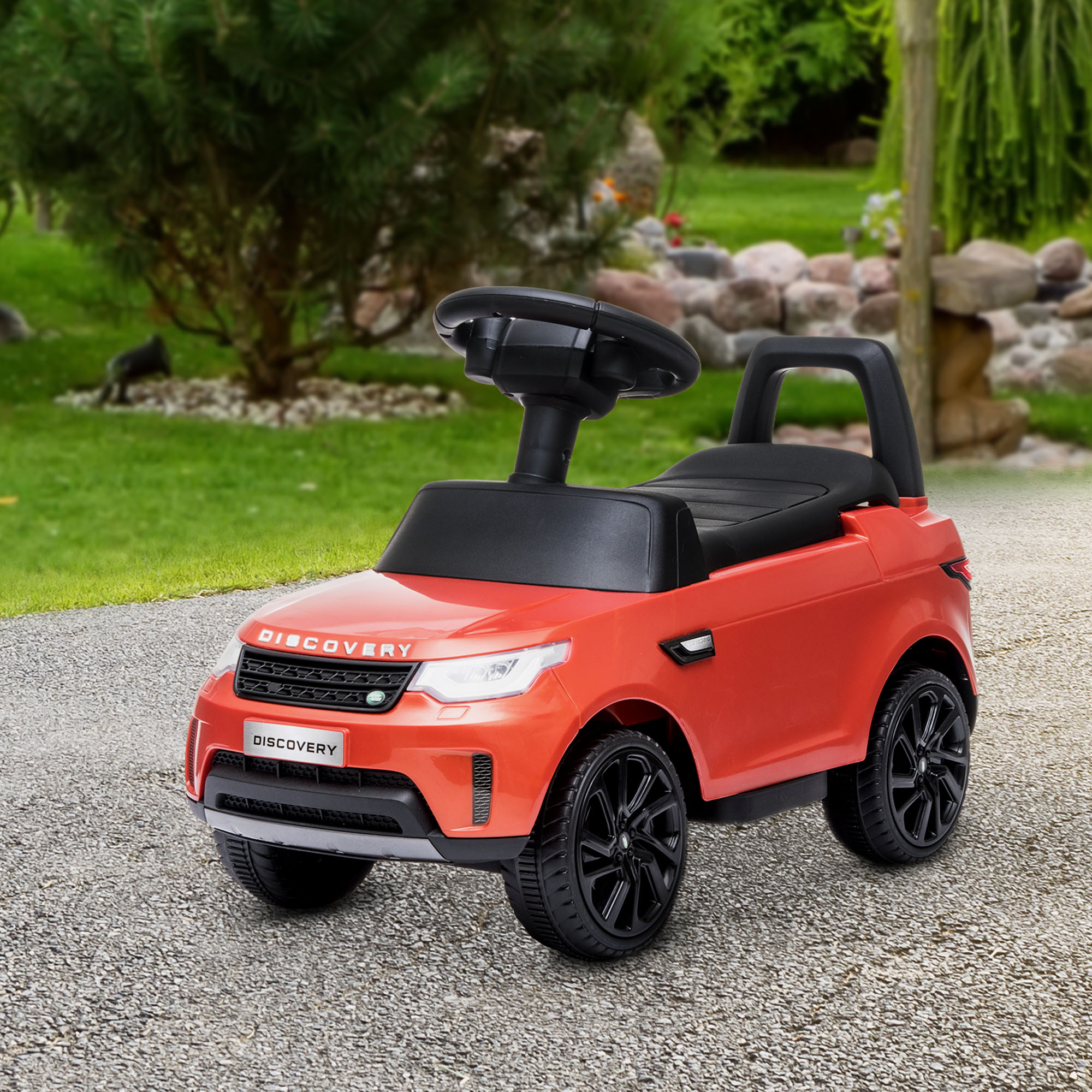 Un'auto giocattolo rossa su una superficie di ghiaia. Un albero e un giardino sono sullo sfondo.