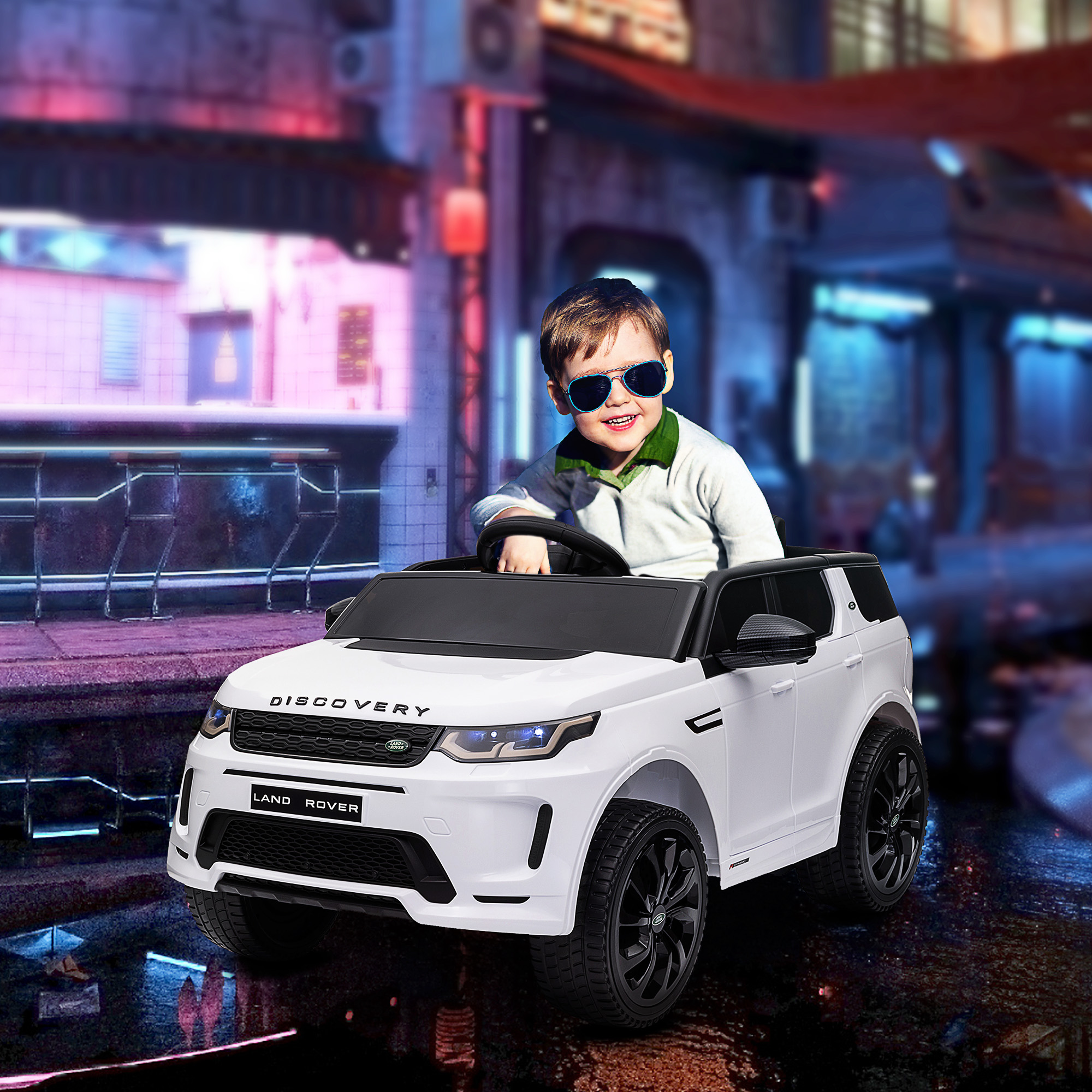 Un niño pequeño con gafas de sol conduce un coche de juguete Land Rover blanco en una calle de la ciudad.