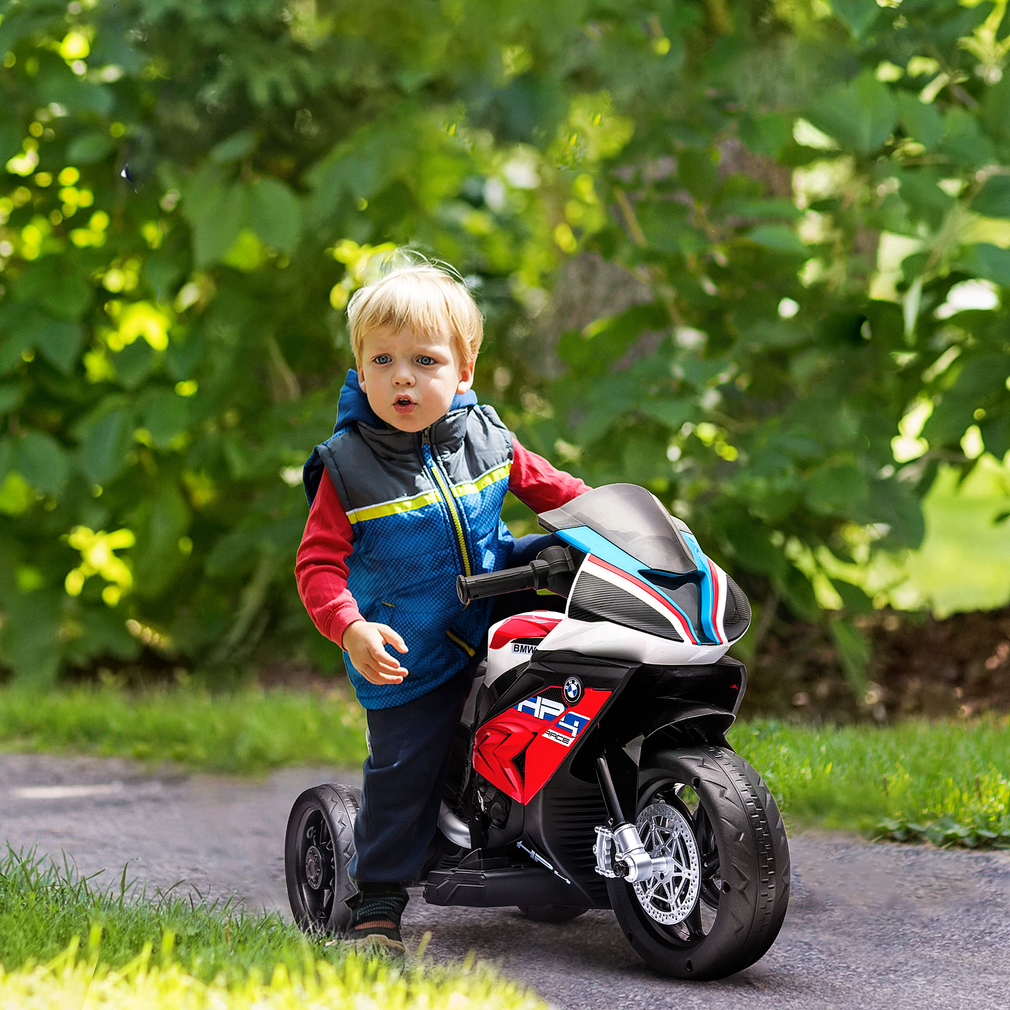 Un bambino biondo è in piedi accanto a una moto giocattolo rossa e nera su un sentiero verde.