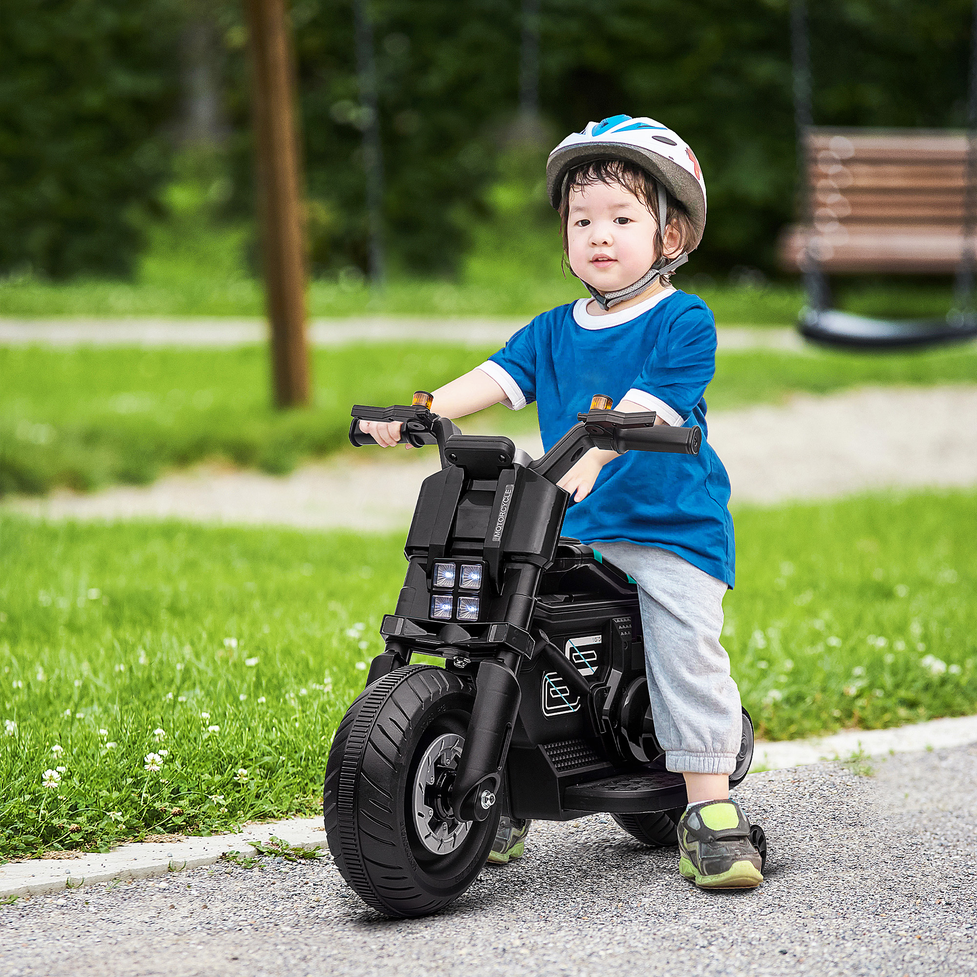 Un bambino piccolo guida una moto giocattolo nera su un sentiero lastricato. Indossa un casco e sorride.