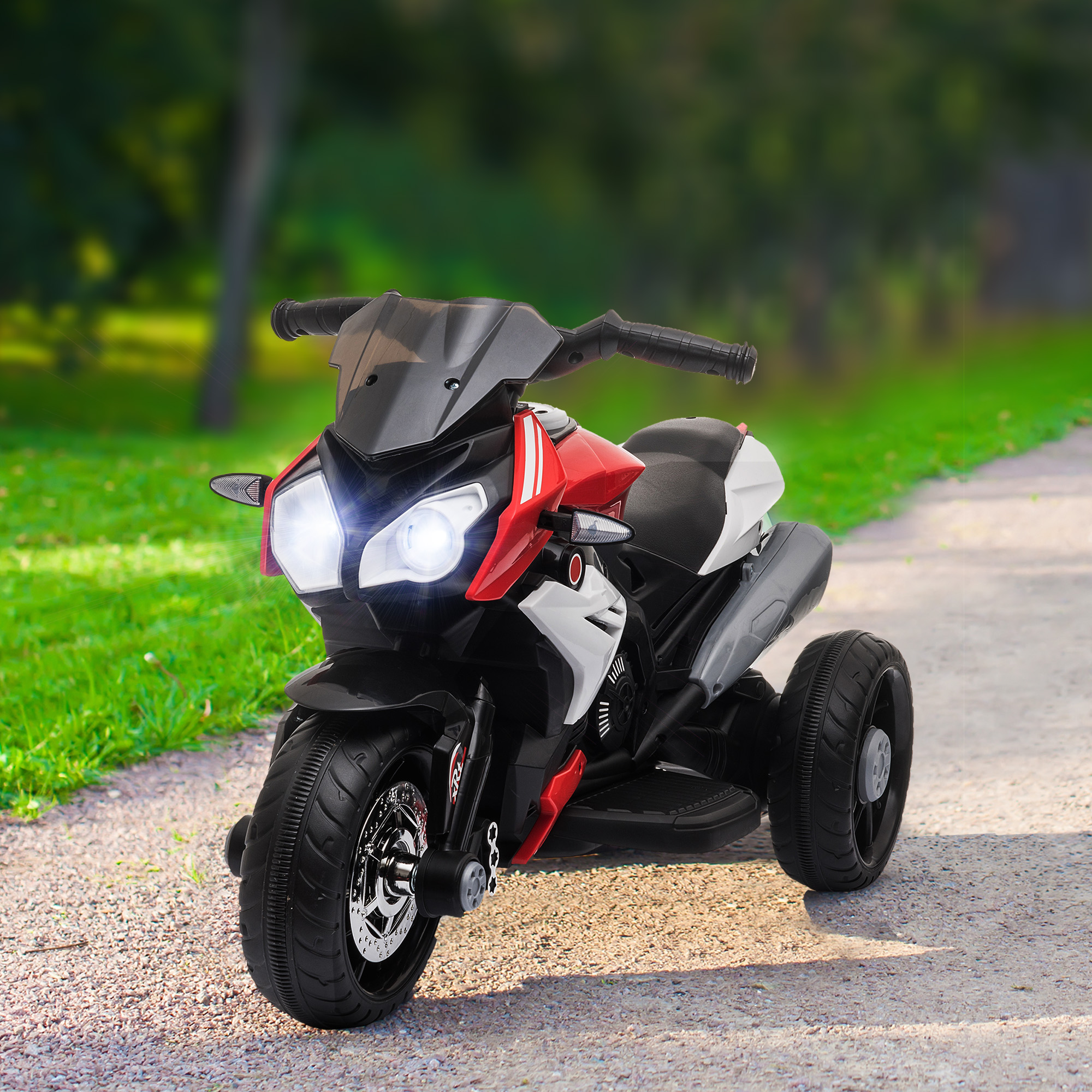 Una moto giocattolo rossa, nera e bianca a tre ruote è parcheggiata su un sentiero pavimentato, circondata dall'erba.