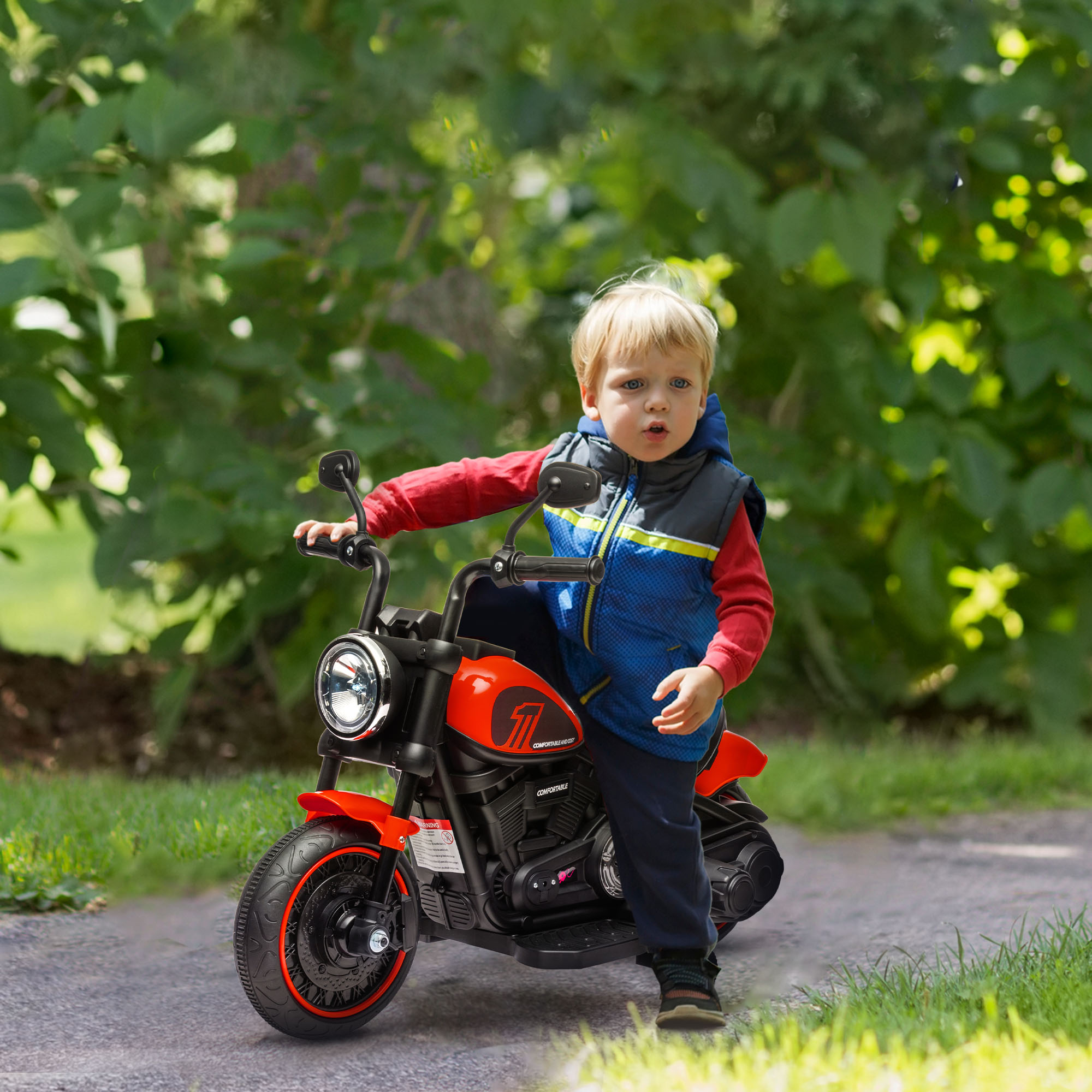 Un ragazzino su una moto giocattolo rossa su una strada asfaltata. Sfondo: alberi ed erba.