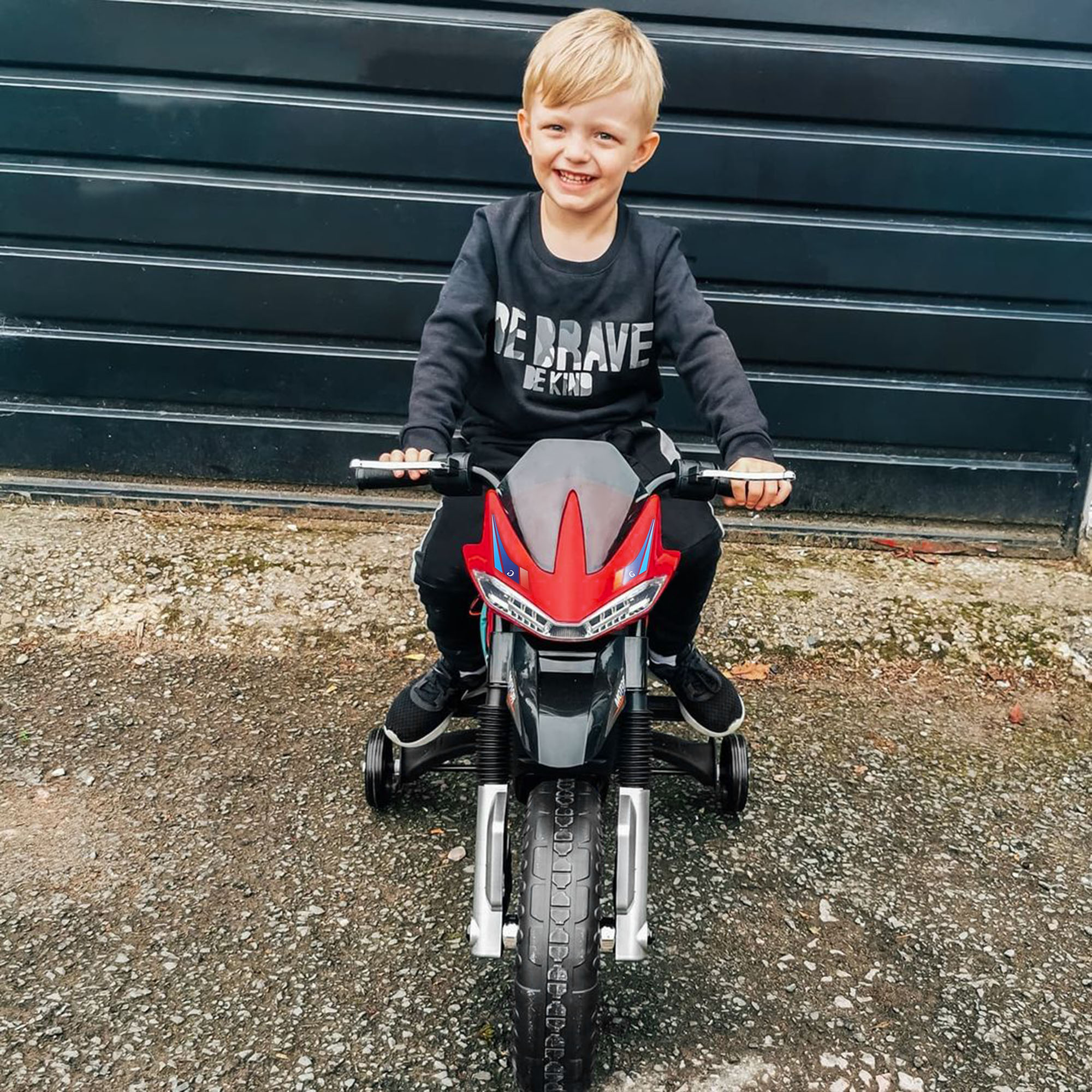 Un bambino biondo sorride, in sella a una moto giocattolo. Top nero con "BE BRAVE".