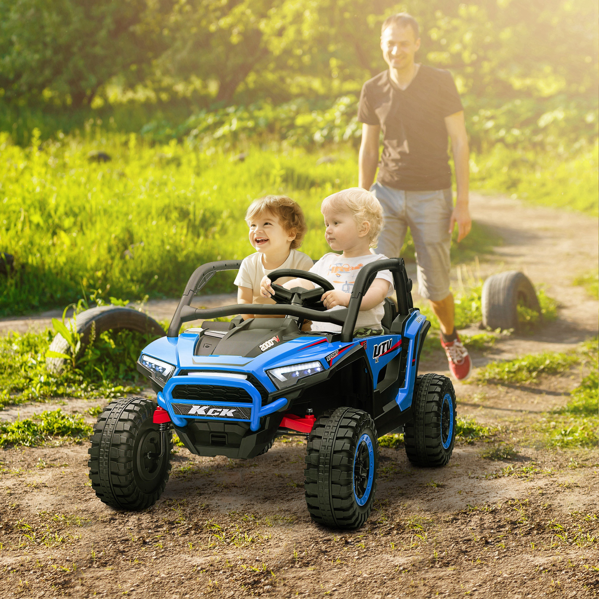 Due bambini in un'auto giocattolo blu, un adulto cammina dietro. L'auto è su un sentiero sterrato con erba verde e alberi.