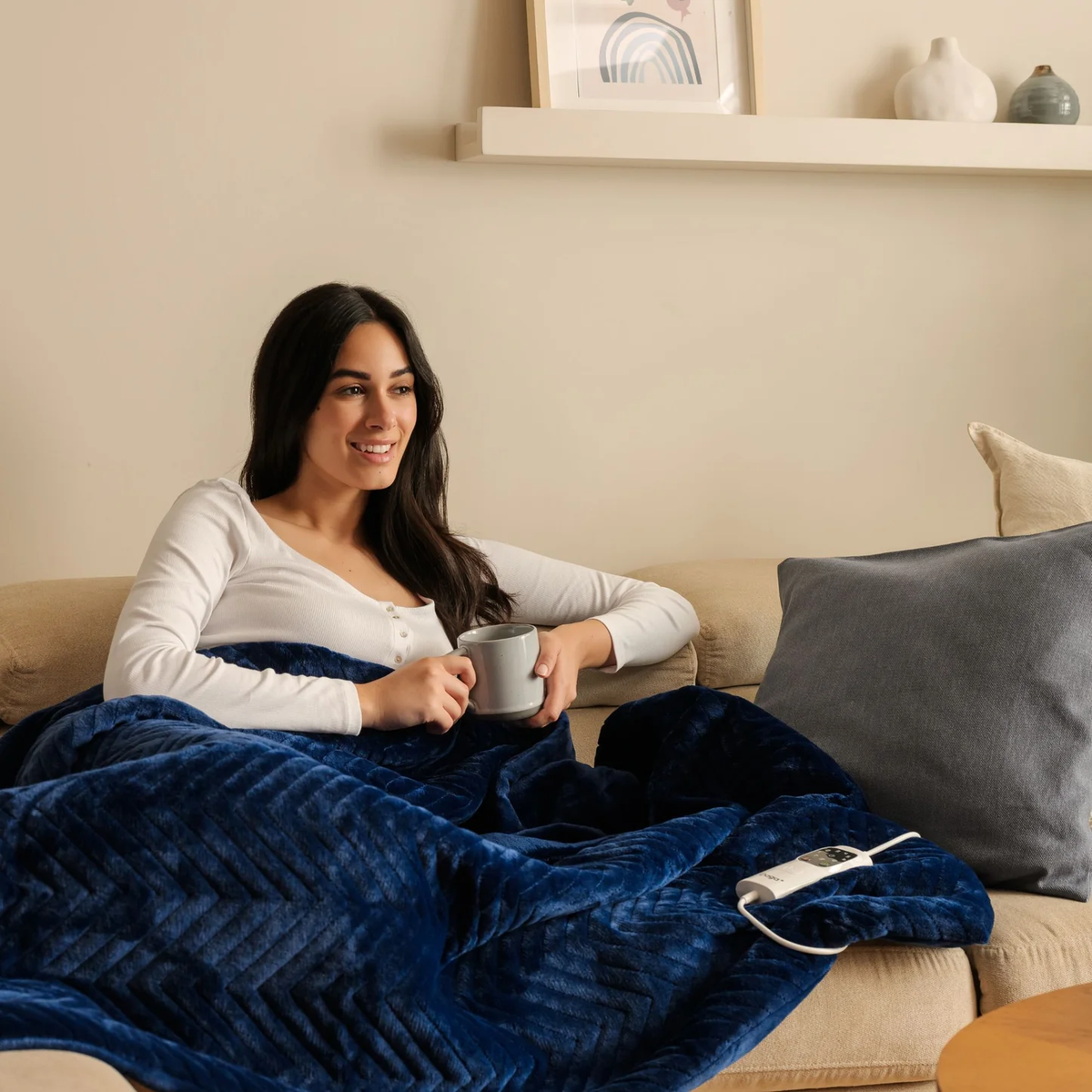 Mujer en sofá bajo una manta azul, sosteniendo una taza.