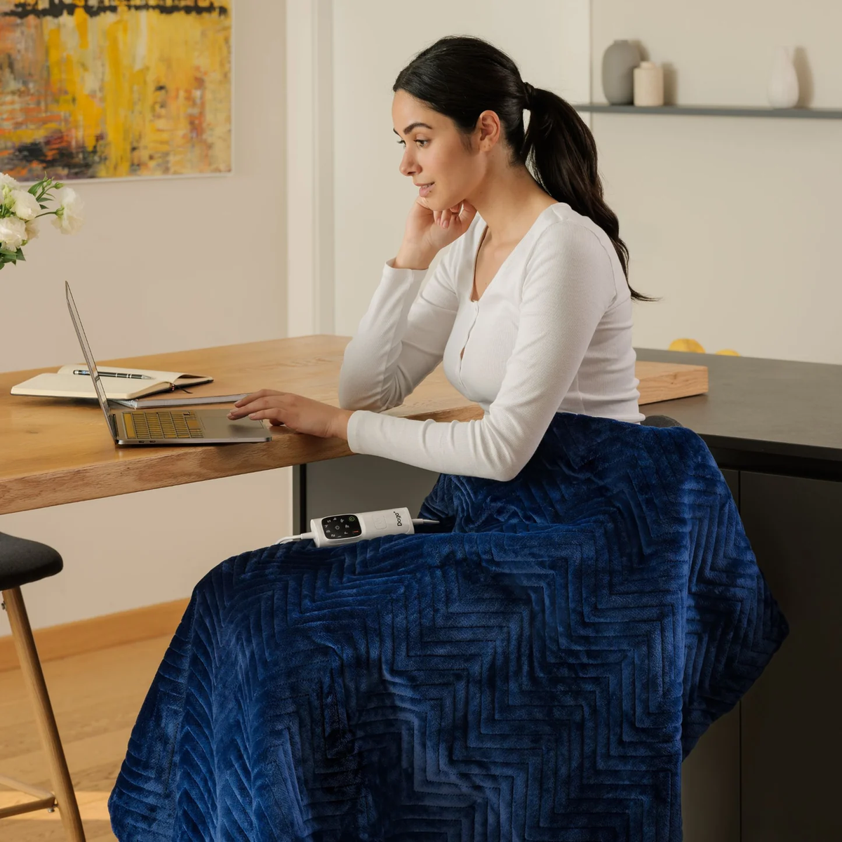 Una mujer sentada en una mesa con un portátil, usando una manta eléctrica azul.