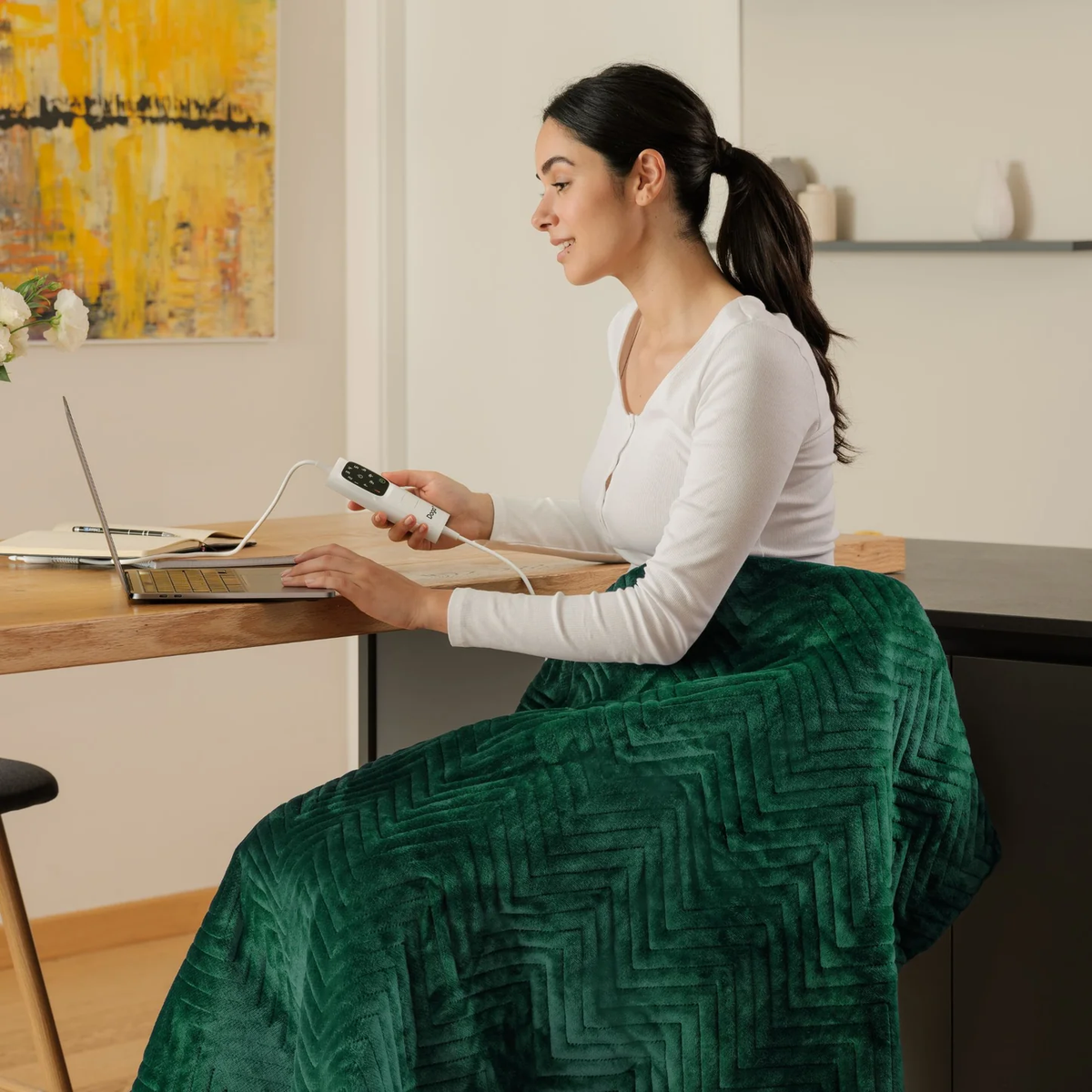 Mujer usando laptop con manta. Manta verde en la silla. Top blanco y cabello oscuro.