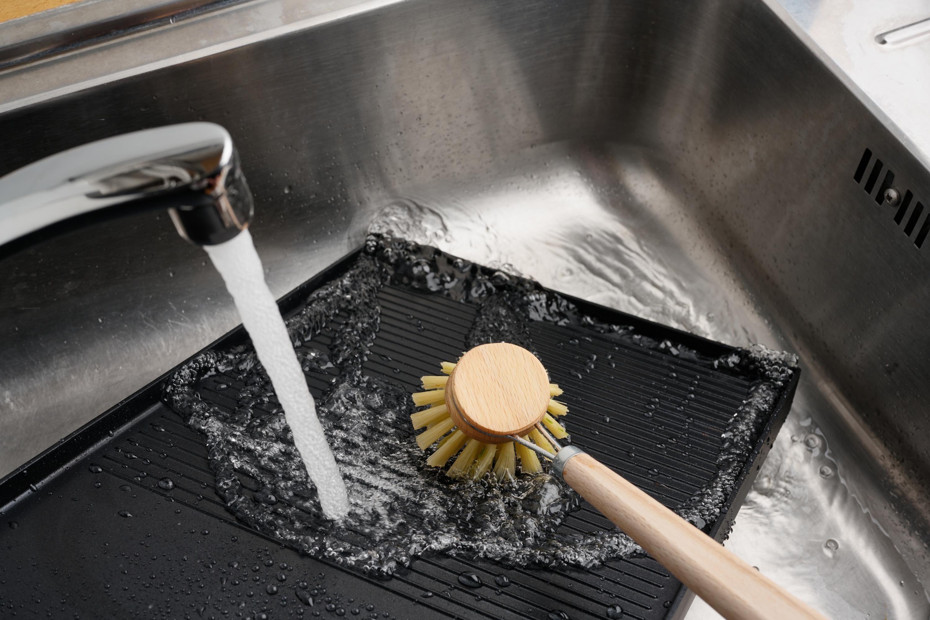 Eine schwarze Grillpfanne wird mit Wasser gewaschen. Eine Holzbürste schrubbt die Pfanne in einem Edelstahlbecken.