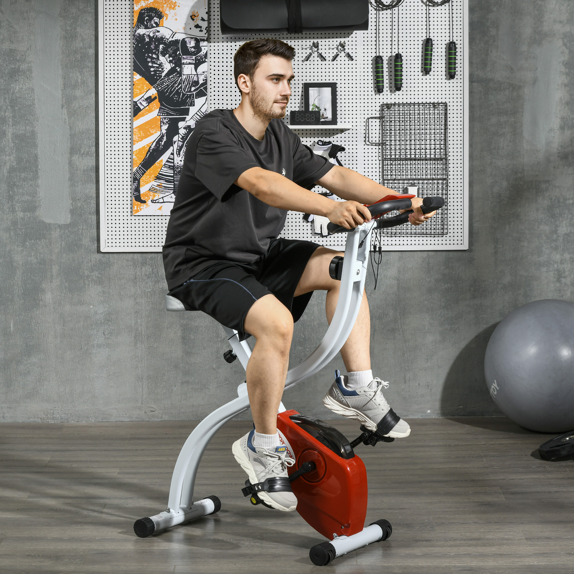 Uomo che usa una cyclette pieghevole. Indossa abiti sportivi. Muro grigio con attrezzature sportive.