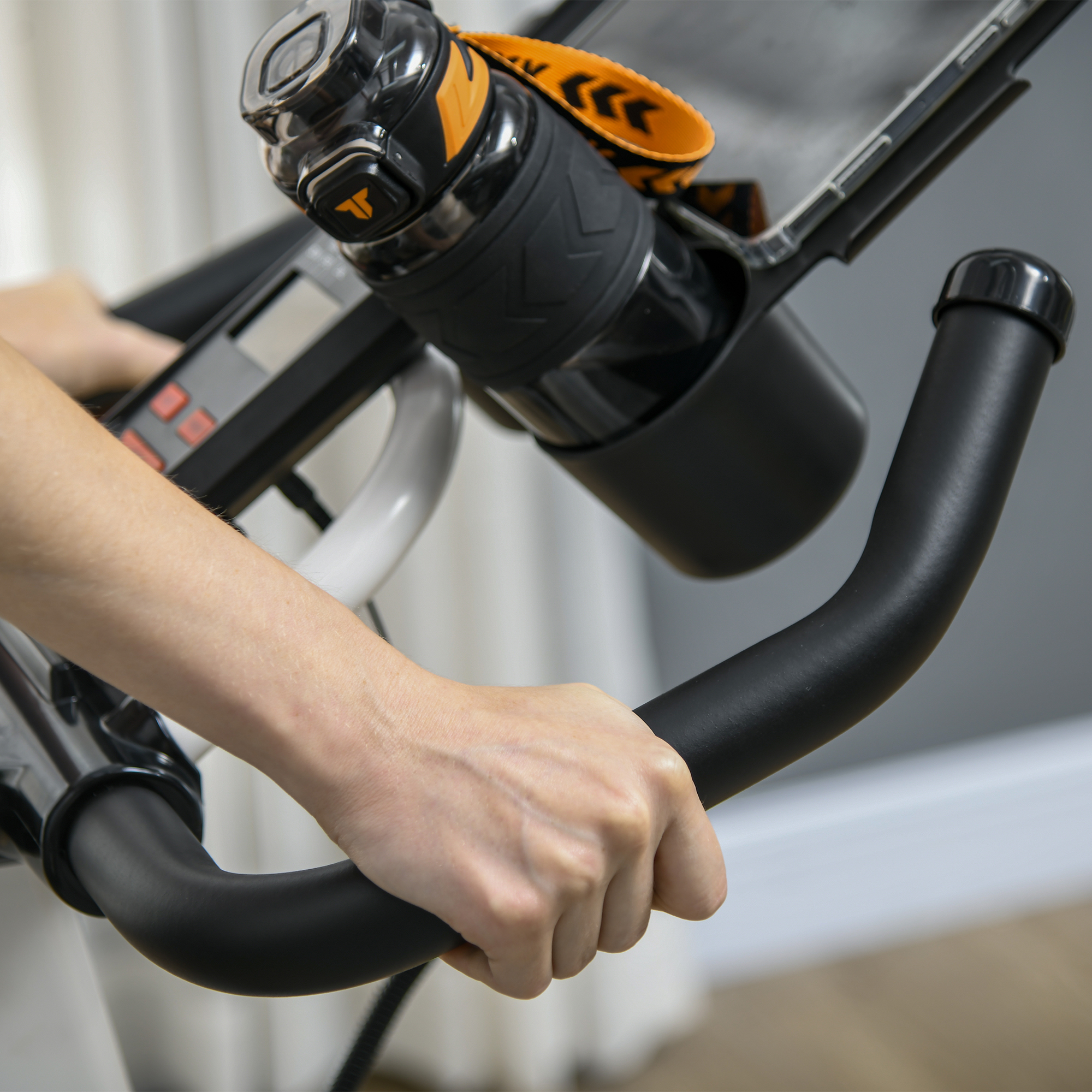 Mano sul manubrio nero della cyclette, bottiglia d'acqua nel supporto. Tenda bianca sullo sfondo.