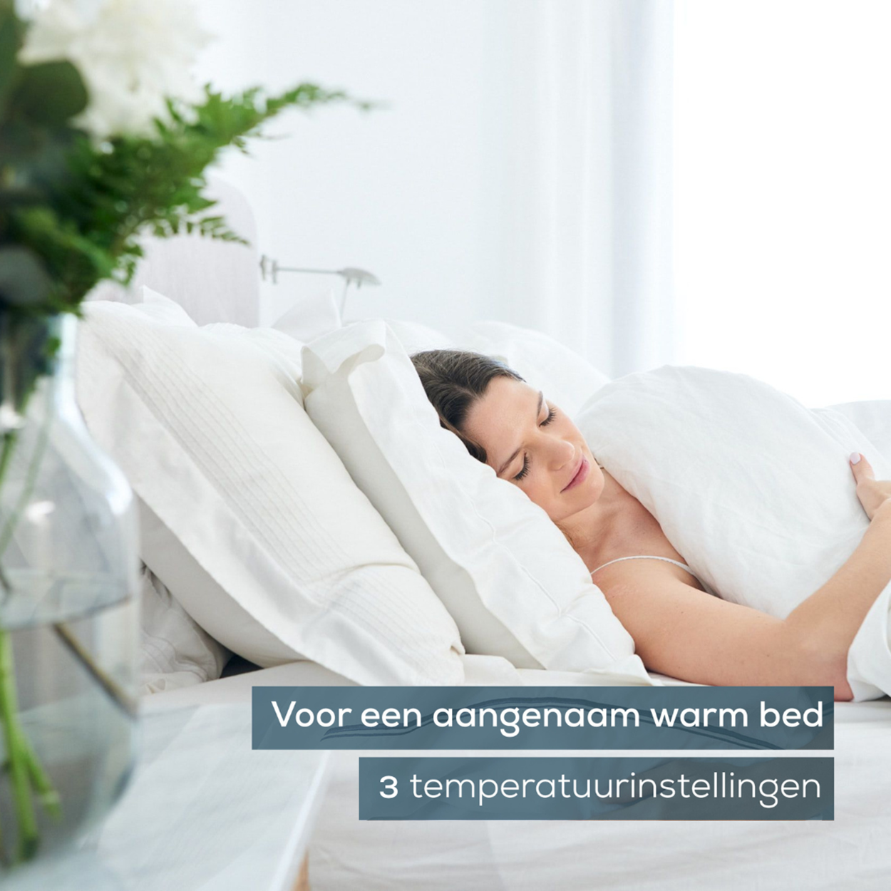 Frau schläft im Bett unter weißen Bettlaken und Kissen, mit Blumen in einer Vase auf einem Nachttisch.