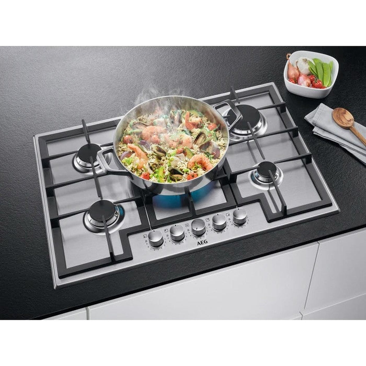 Un piano cottura in acciaio inossidabile con quattro fuochi. Una padella con cibo fuma sui fuochi. Le verdure sono nelle vicinanze.
