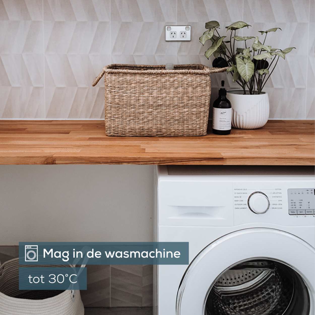 Eine Waschmaschine und ein Wäschekorb befinden sich in einem Raum. Der Text lautet: 'Mag in de wasmachine tot 30°C'.