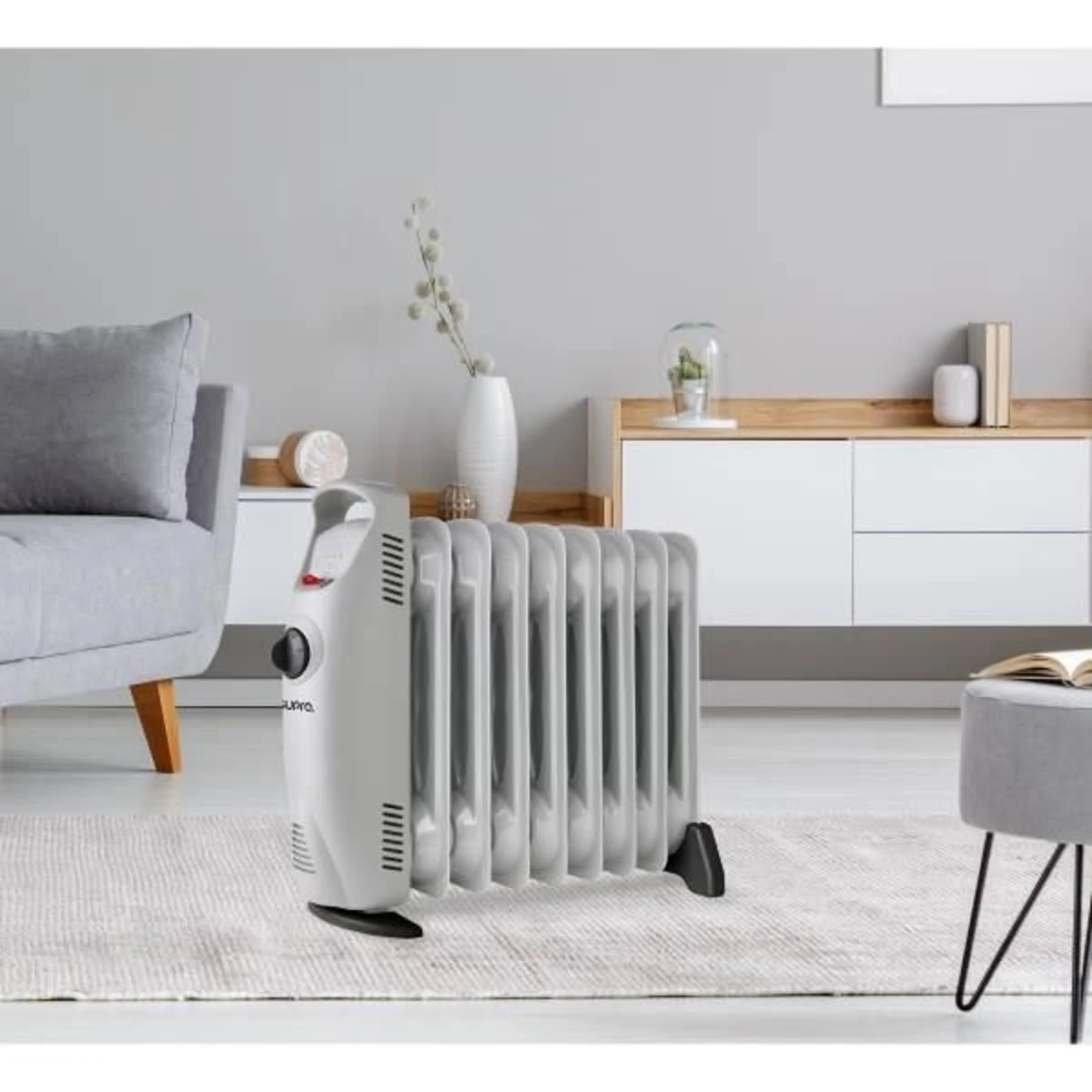 Szary grzejnik elektryczny stoi na dywanie w pobliżu sofy i wazonu. Biała szafka w tle.
