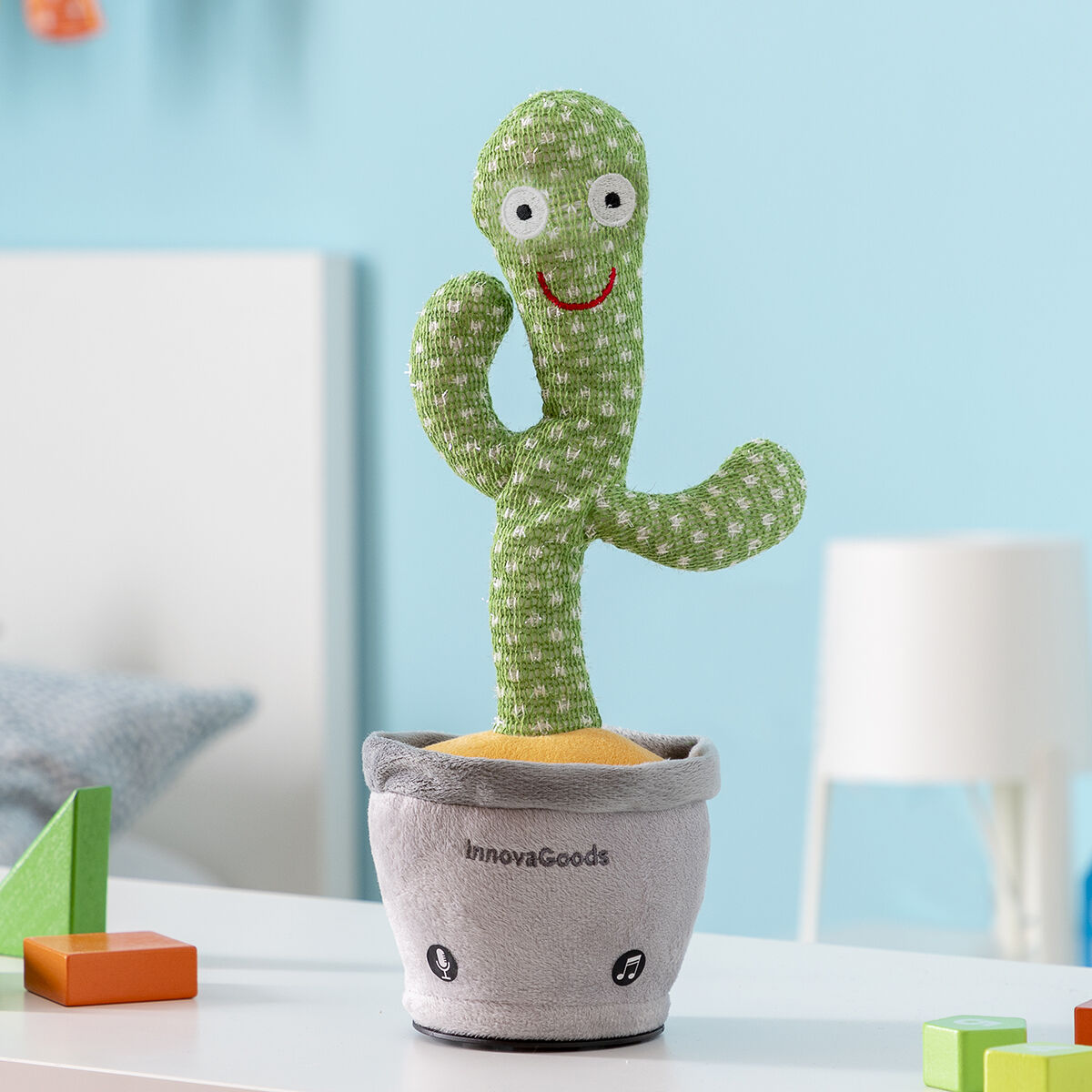 Un giocattolo a forma di cactus verde in un vaso grigio su un tavolo bianco. Sullo sfondo un muro blu.
