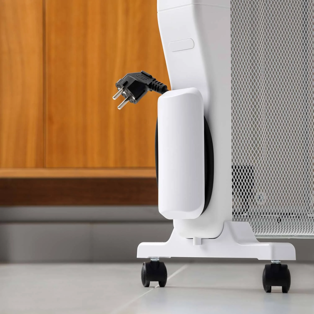 Biały grzejnik elektryczny z nawijarką i wtyczką. Ma kółka i stoi obok drewnianej ściany.