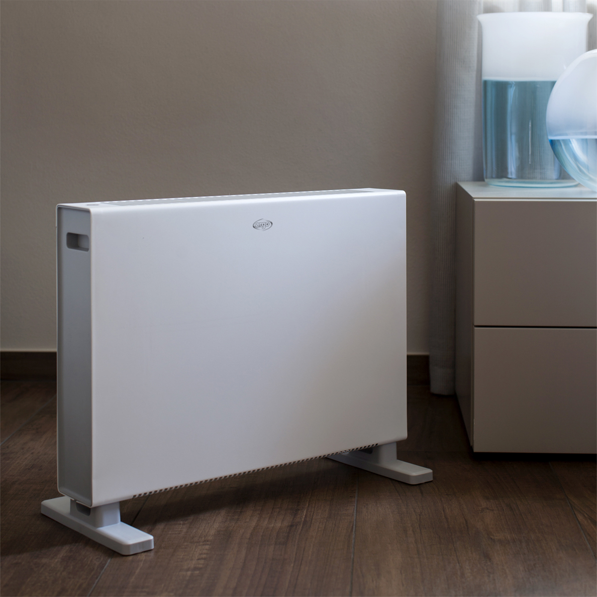 Biały grzejnik na drewnianej podłodze, przed ścianą. Po prawej szafka i wazon.