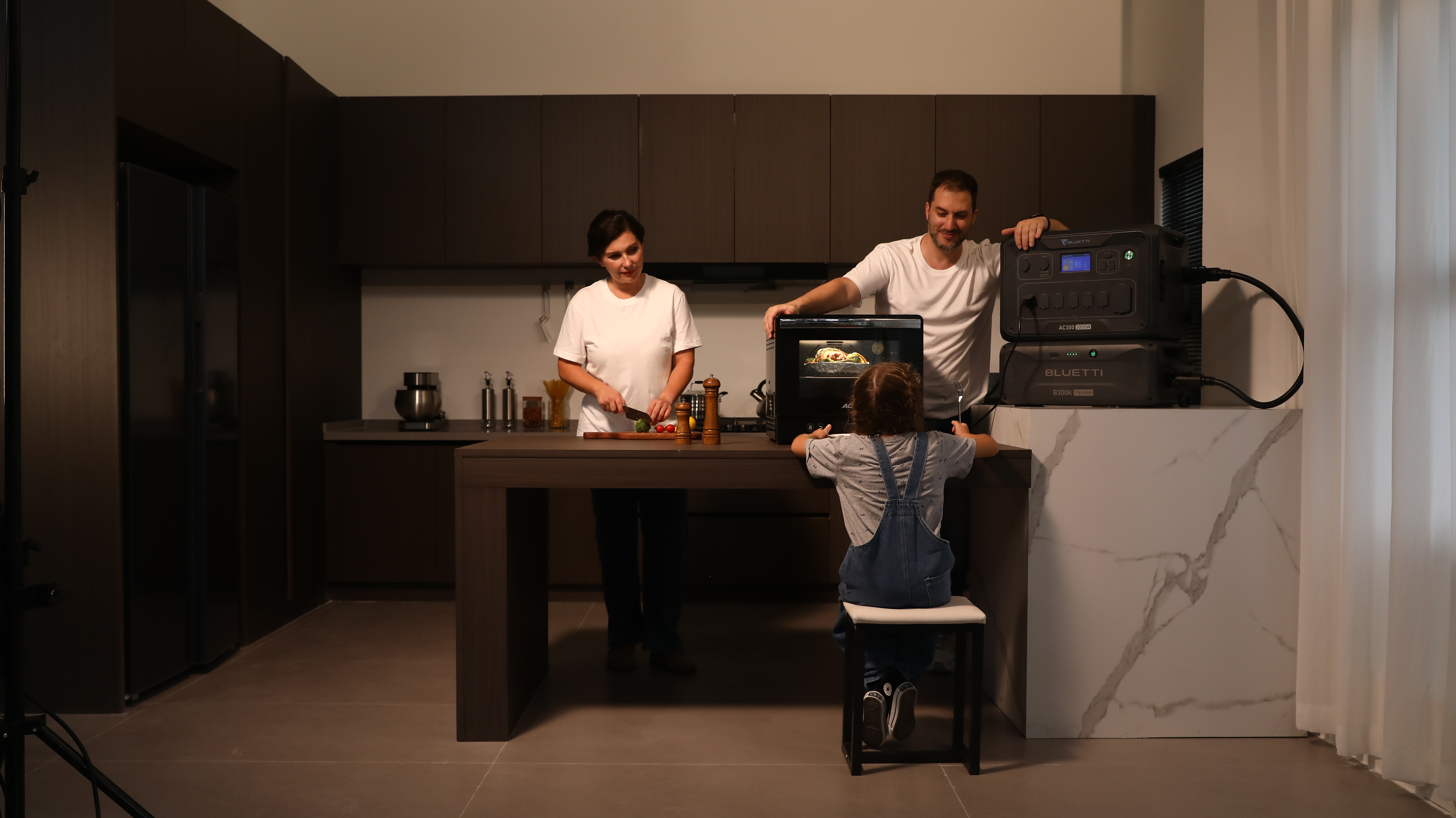 Rodzina gotuje w kuchni. Dziecko siedzi przy stole. Jednostki zasilania widoczne w tle.