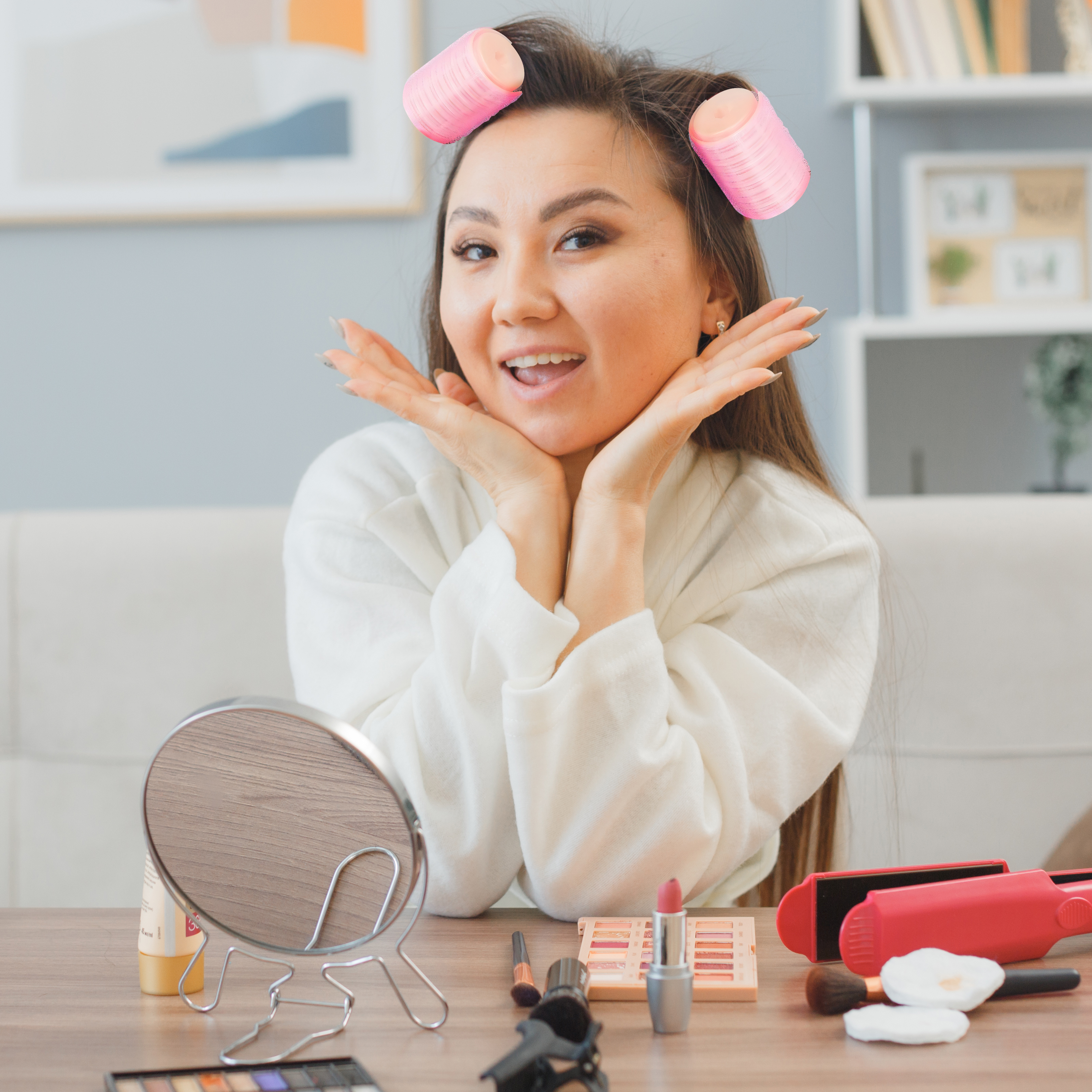 Eine Frau mit Lockenwicklern im Haar lächelt in die Kamera, umgeben von Make-up. Hintergrund: Wohnzimmer.