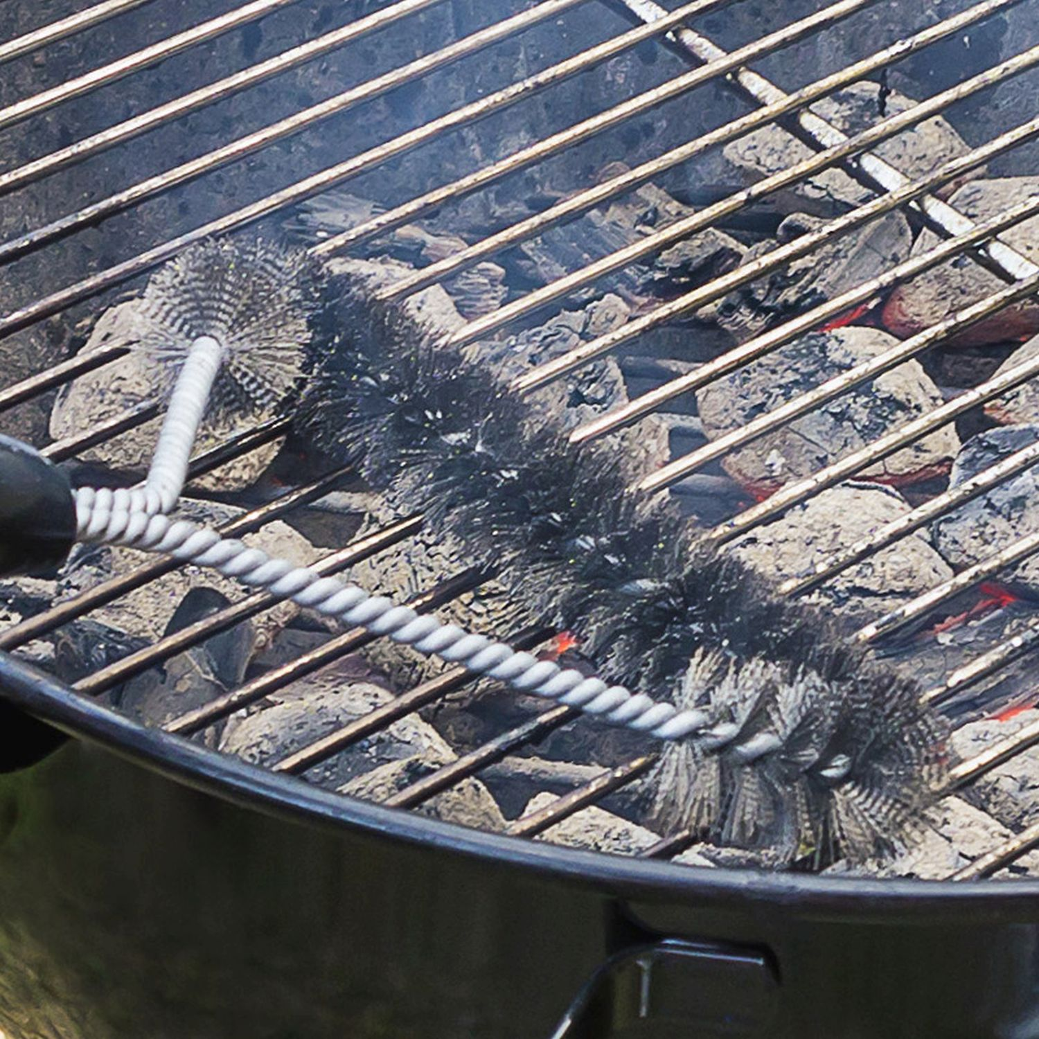 Nahaufnahme einer Grillbürste, die einen Grill reinigt, mit verkohlter Holzkohle darunter. Rauch steigt von der Holzkohle auf.