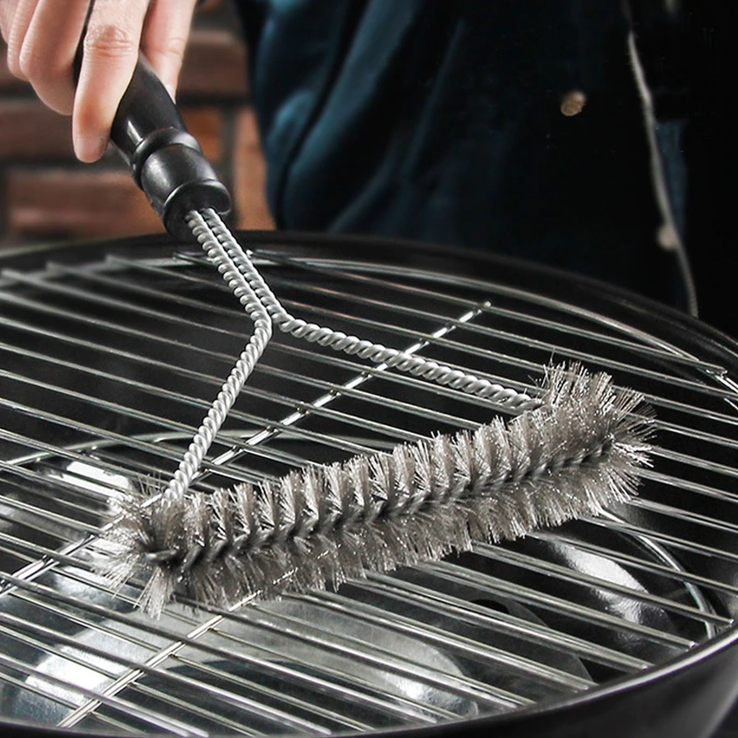 Eine Edelstahl-Grillbürste reinigt einen schwarzen Grillrost. Eine Person hält die Bürste.