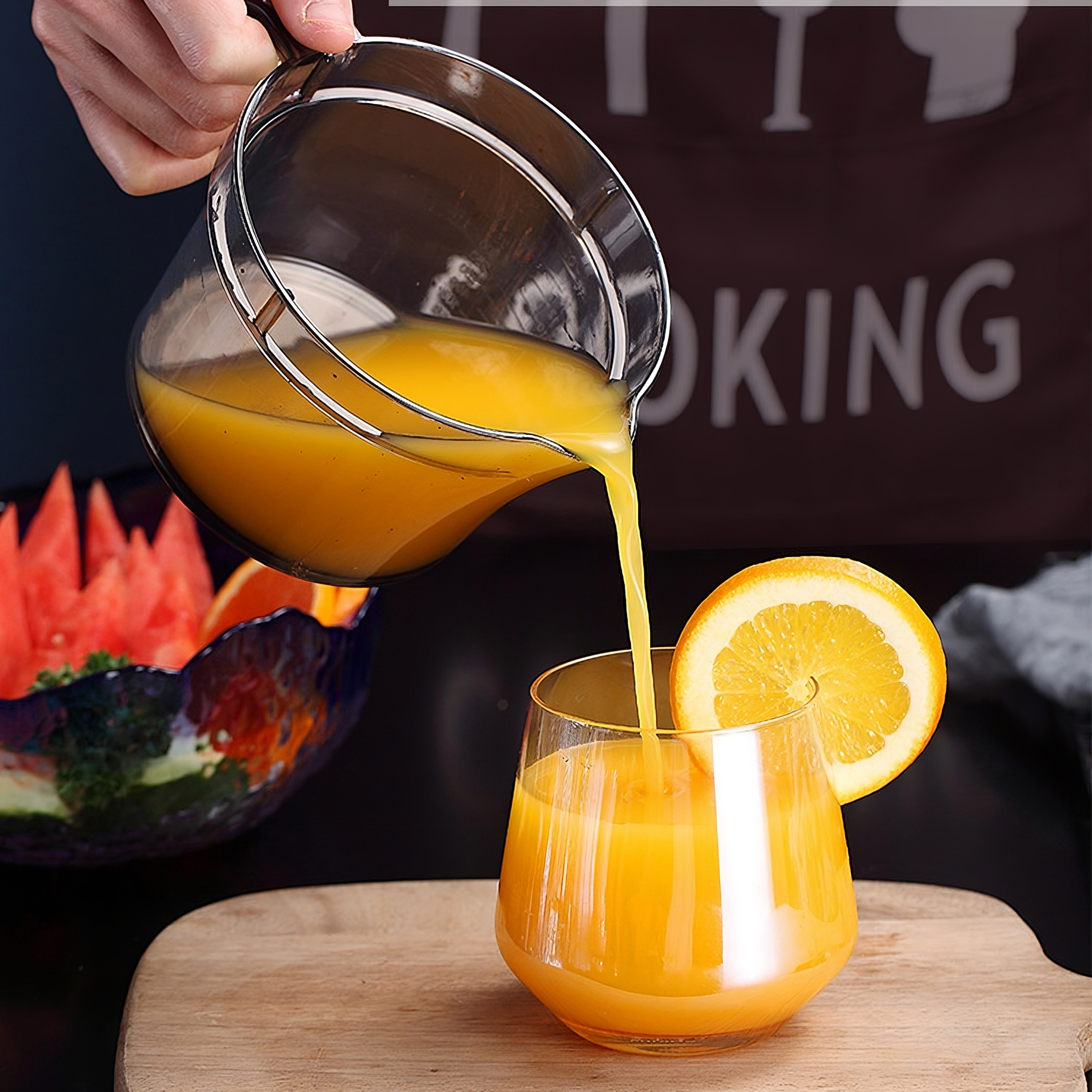 Eine Person gießt Orangensaft in ein Glas mit einer Orangenscheibe. Ein Holzbrett im Vordergrund.