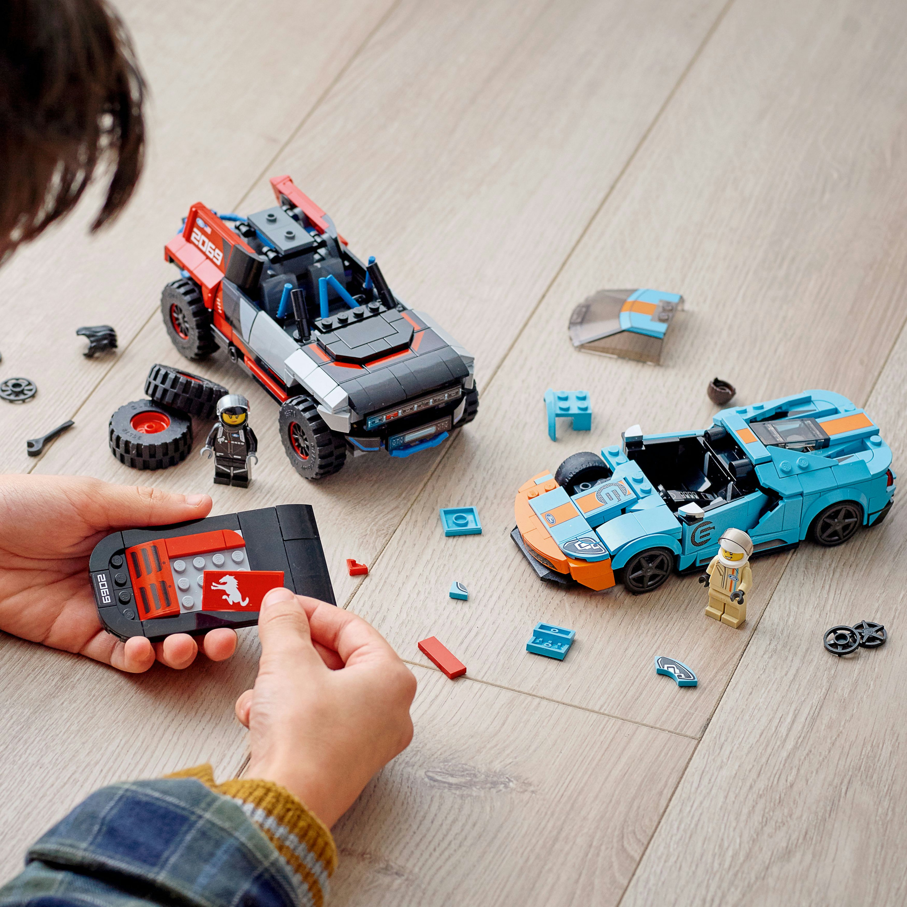 Osoba bawiąca się samochodami Lego i częściami samochodowymi, z dwoma samochodami na drewnianej powierzchni.