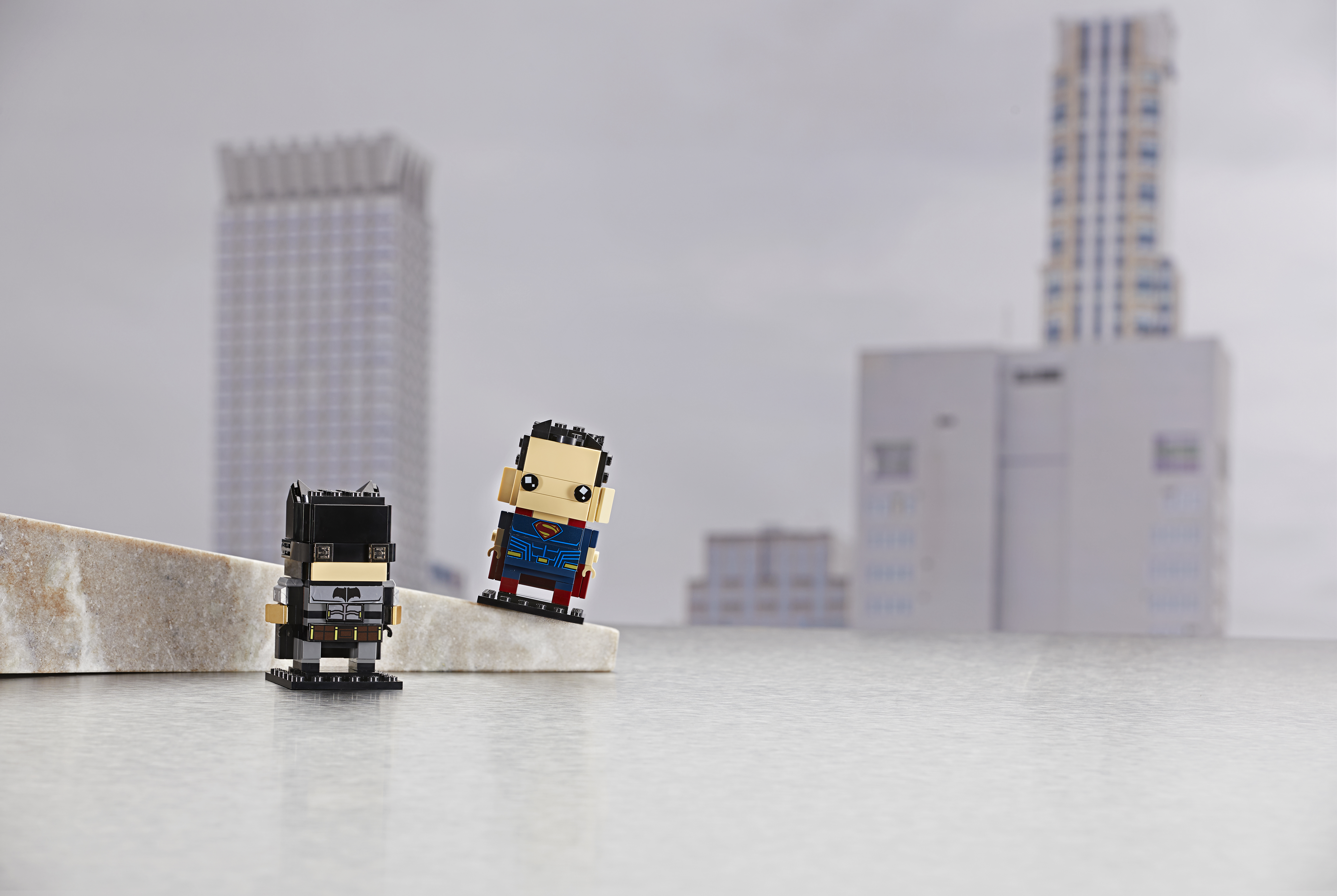 Lego Batman en Superman brickheadz figuren op een oppervlak, met stadsachtergrond.