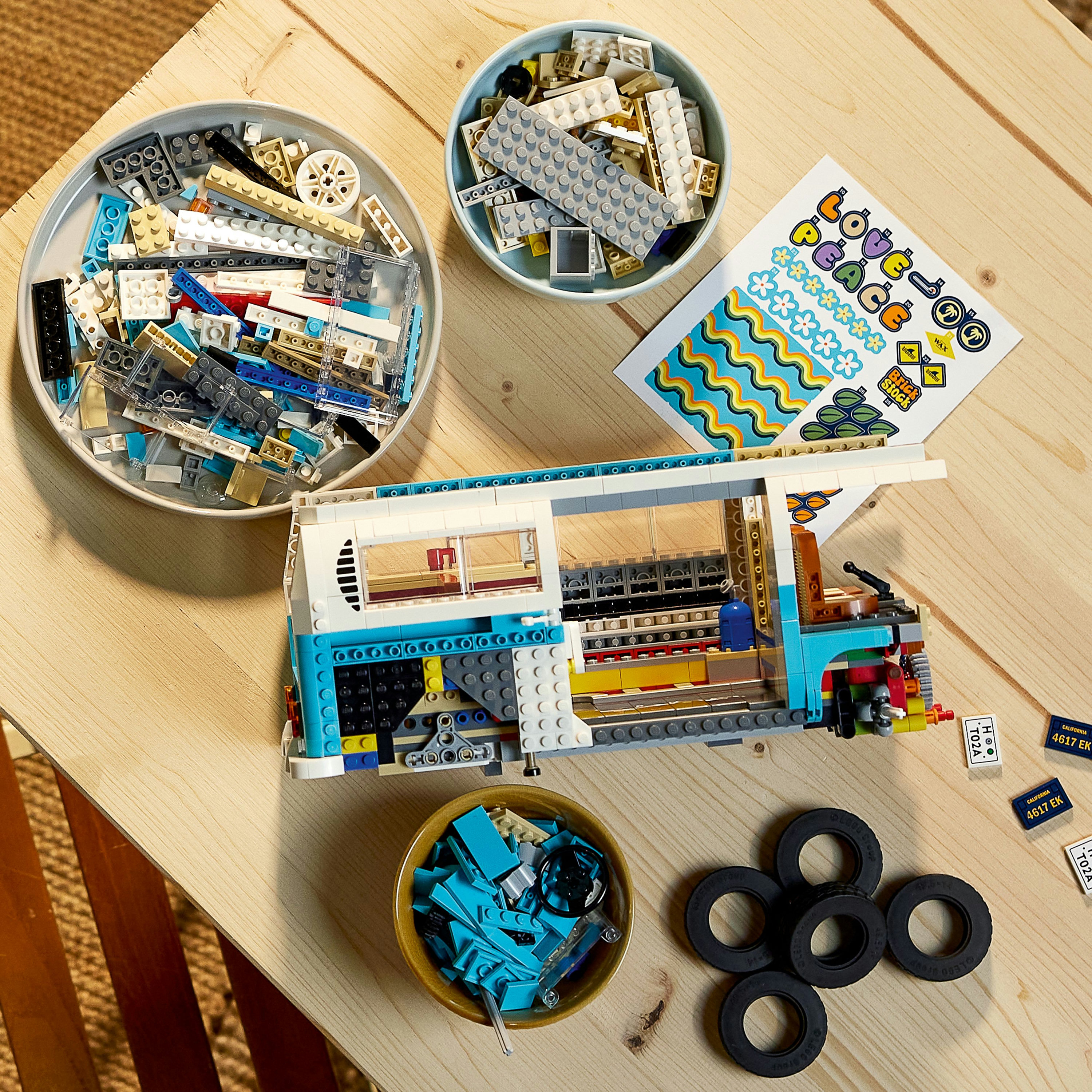Lego campervan in aanbouw met diverse Lego-stenen en stickers op een houten tafel.