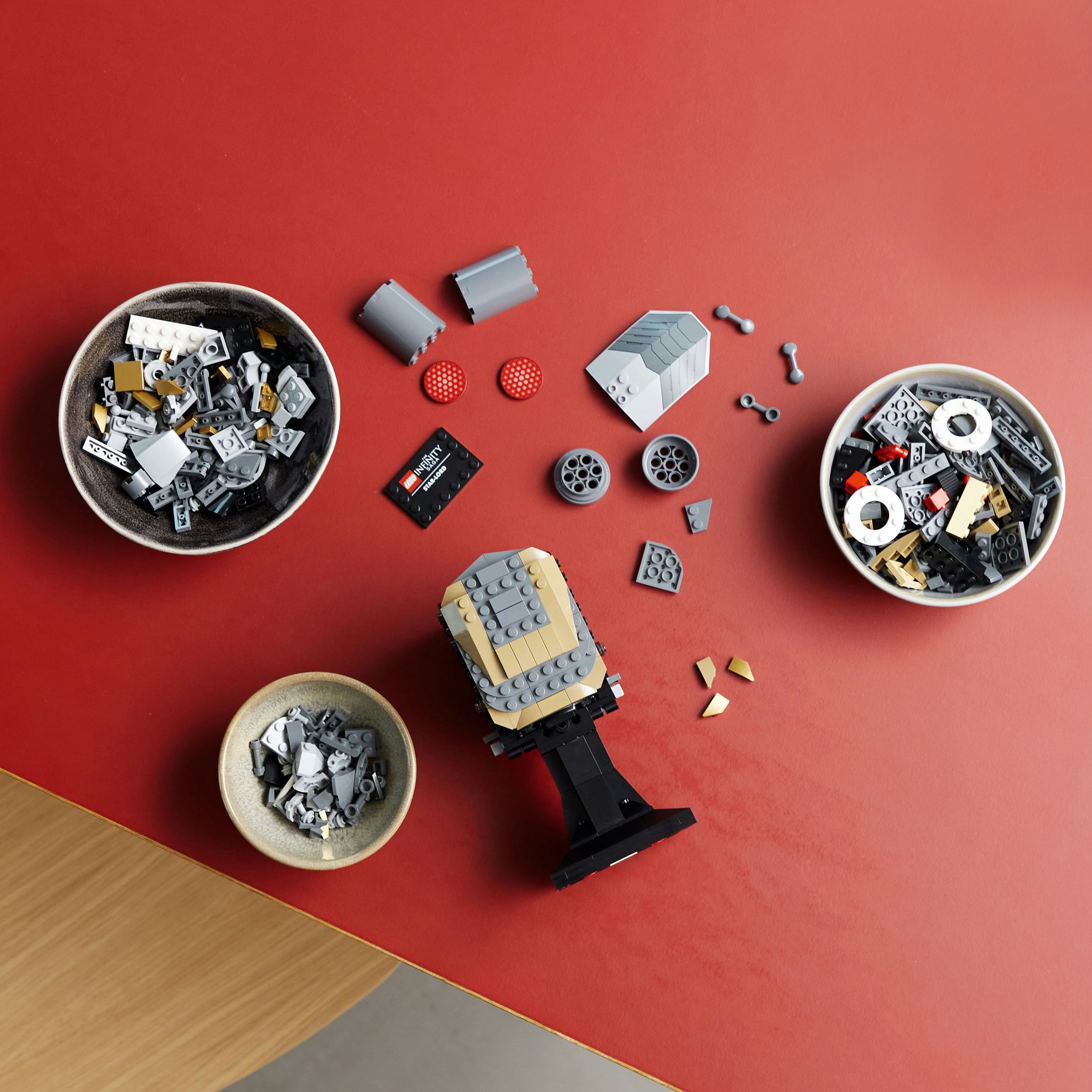 Klocki Lego w miskach na czerwonej powierzchni. Obecne są różne szare, czarne i złote klocki oraz model Lego.
