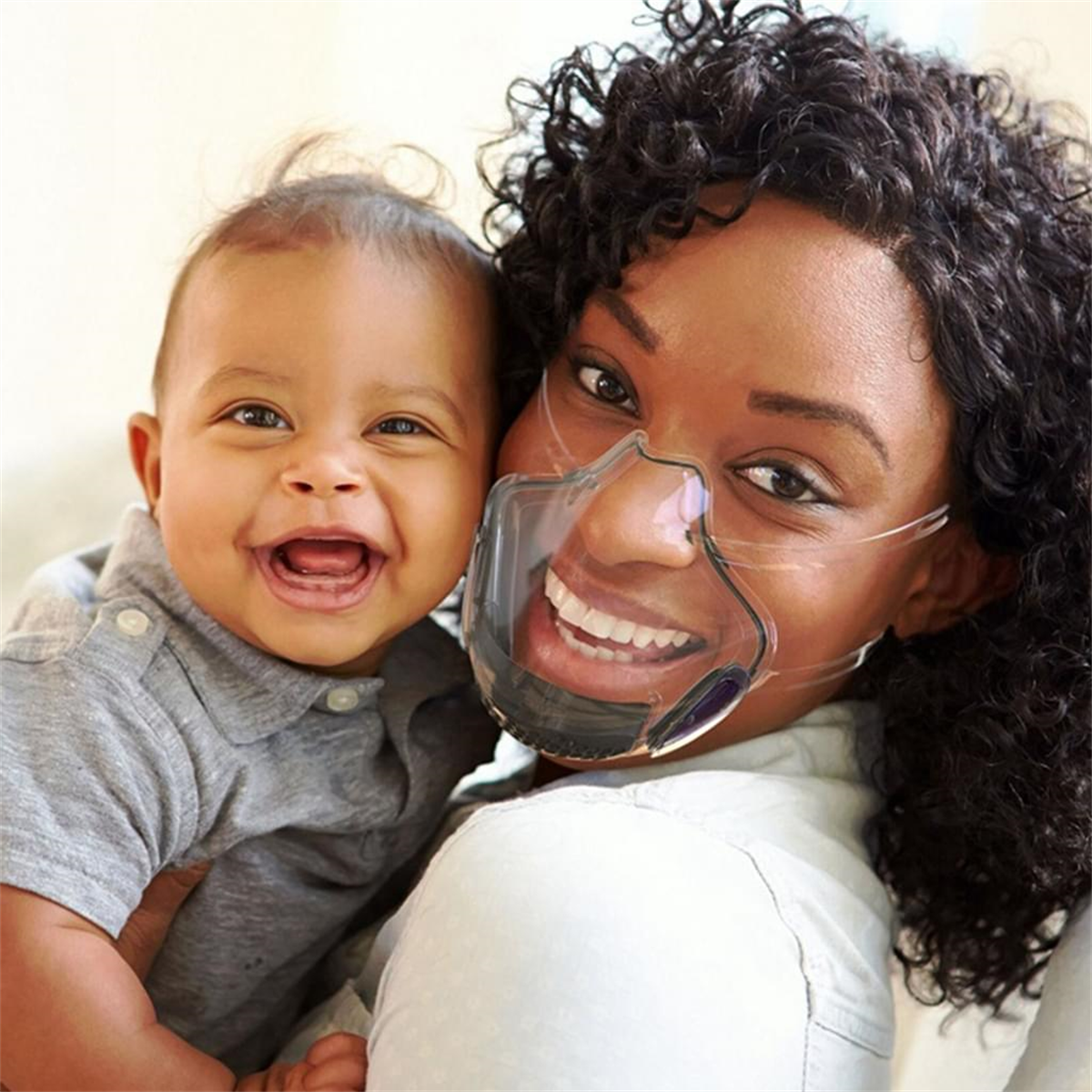 Frau und Baby lächeln, Frau trägt eine transparente Maske. Der Hintergrund ist hell.