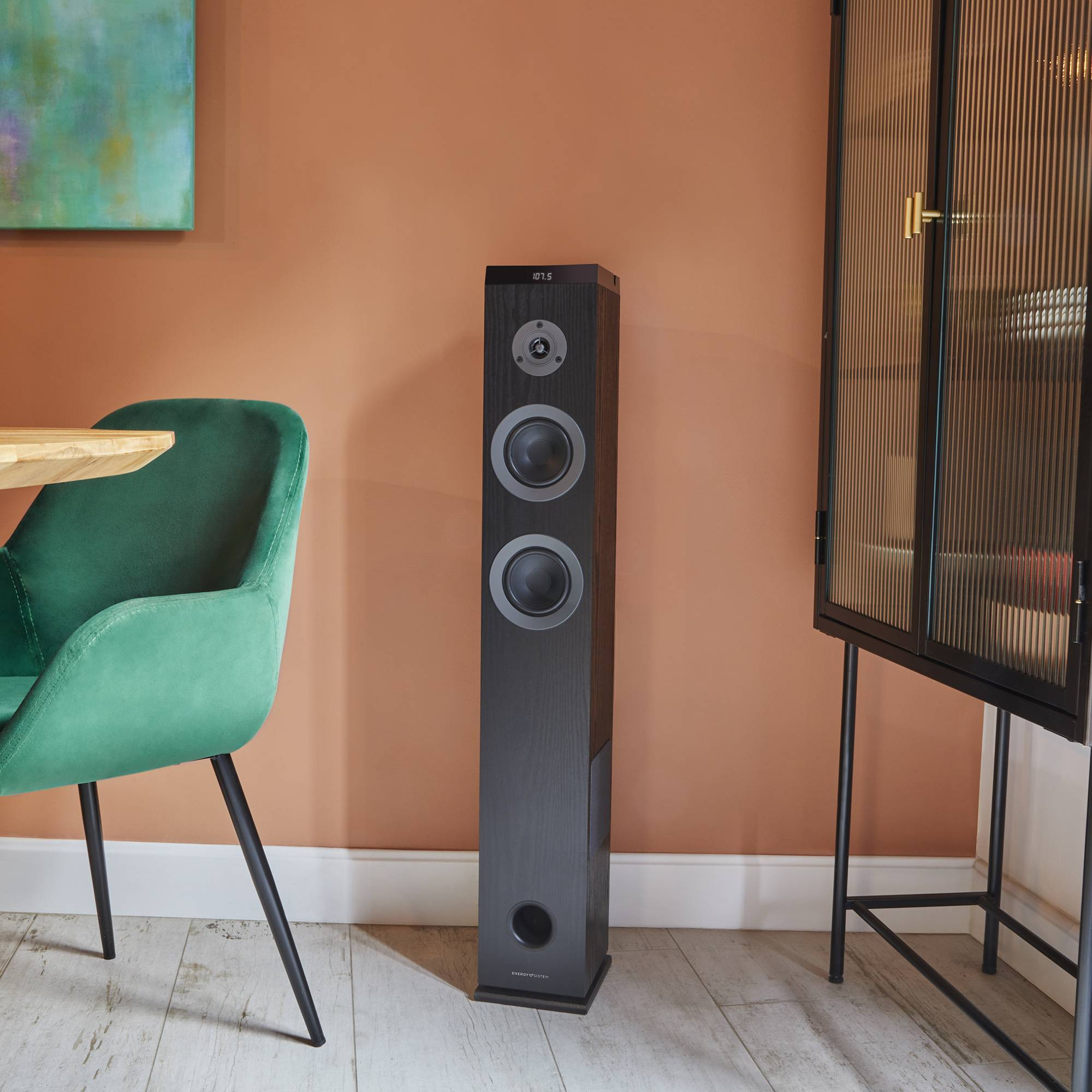 Un altavoz negro alto junto a un mueble y una silla verde. La pared es naranja.