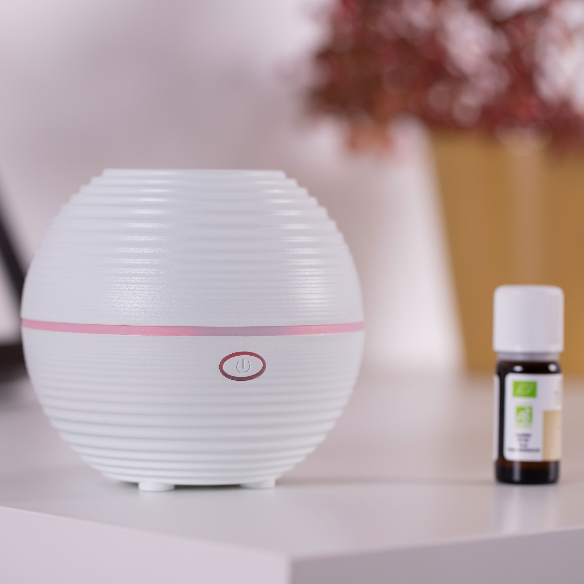 A white, round essential oil diffuser with a pink button and a small oil bottle.