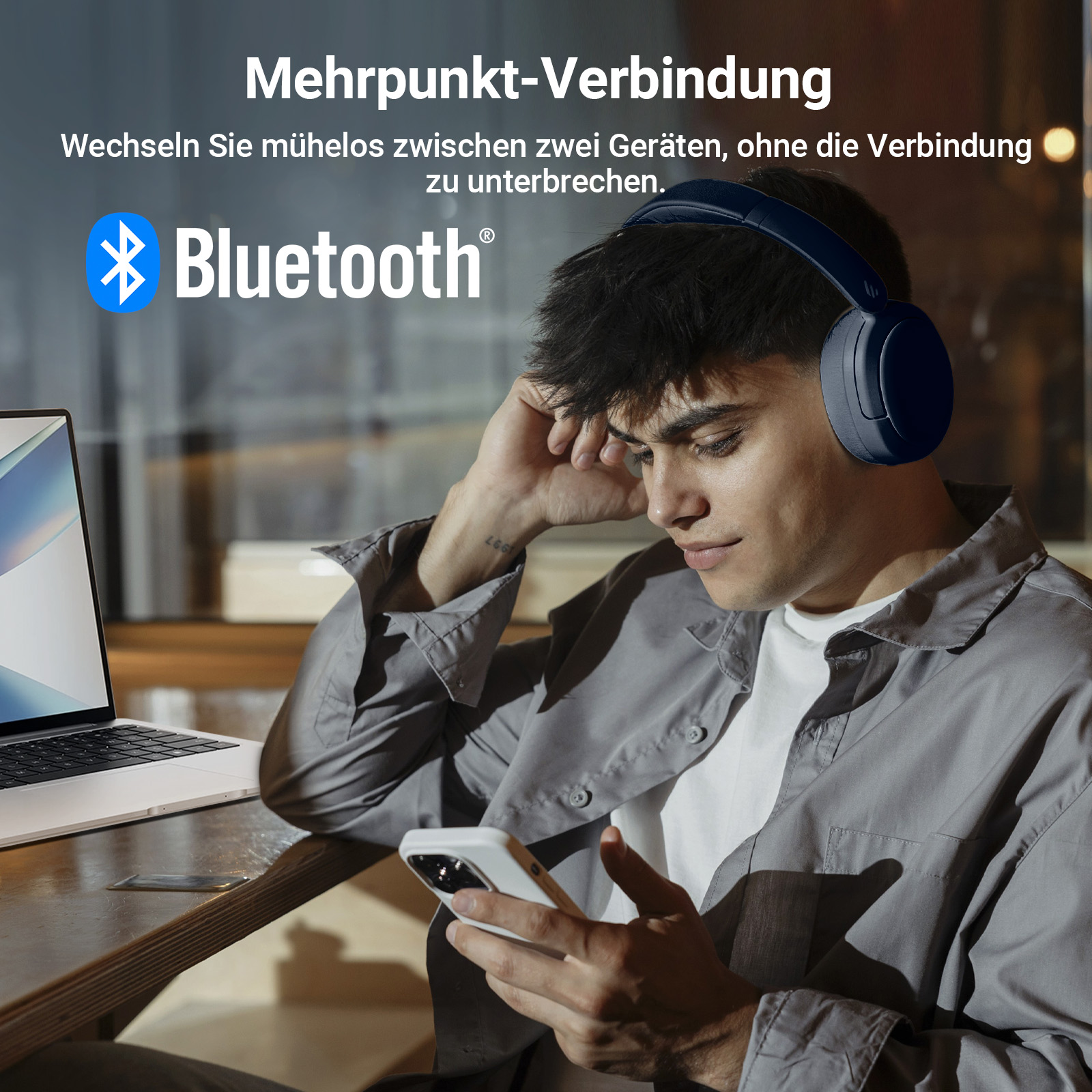 Ein Mann schaut auf sein Telefon, Bluetooth-Kopfhörer auf, und ein Laptop im Hintergrund.