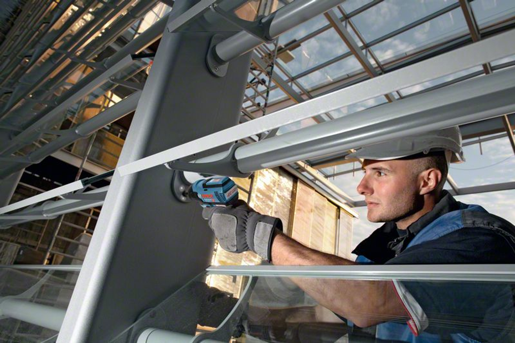 Mężczyzna w kasku używa wiertarki na metalowej konstrukcji. Tło to niebo.