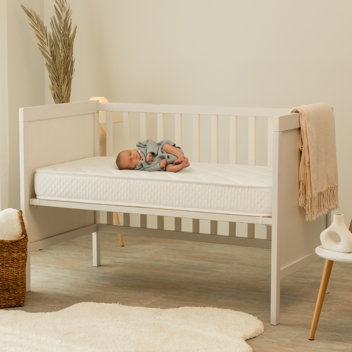 Un bebé durmiendo en una cuna blanca con una manta beige y una alfombra blanca.