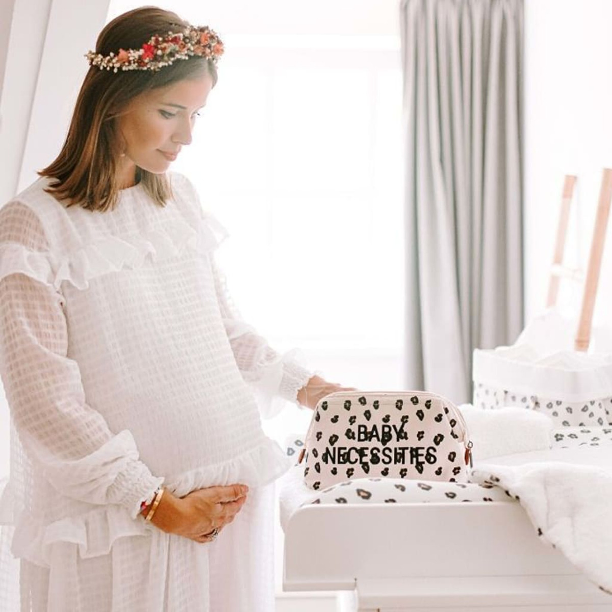 Schwangere Frau in weißem Kleid hält ihren Bauch. Blumengirlande und eine Baby-Utensilientasche sind in der Nähe.