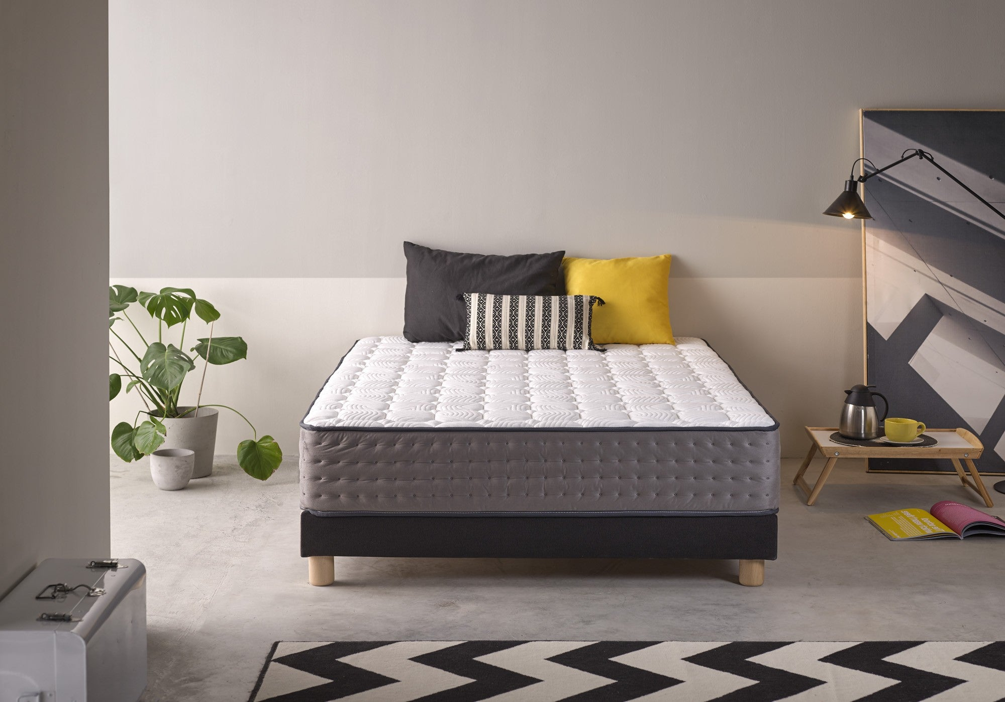 Escena de dormitorio con cama, almohadas, lámpara y mesita de noche. Plantas y alfombra.