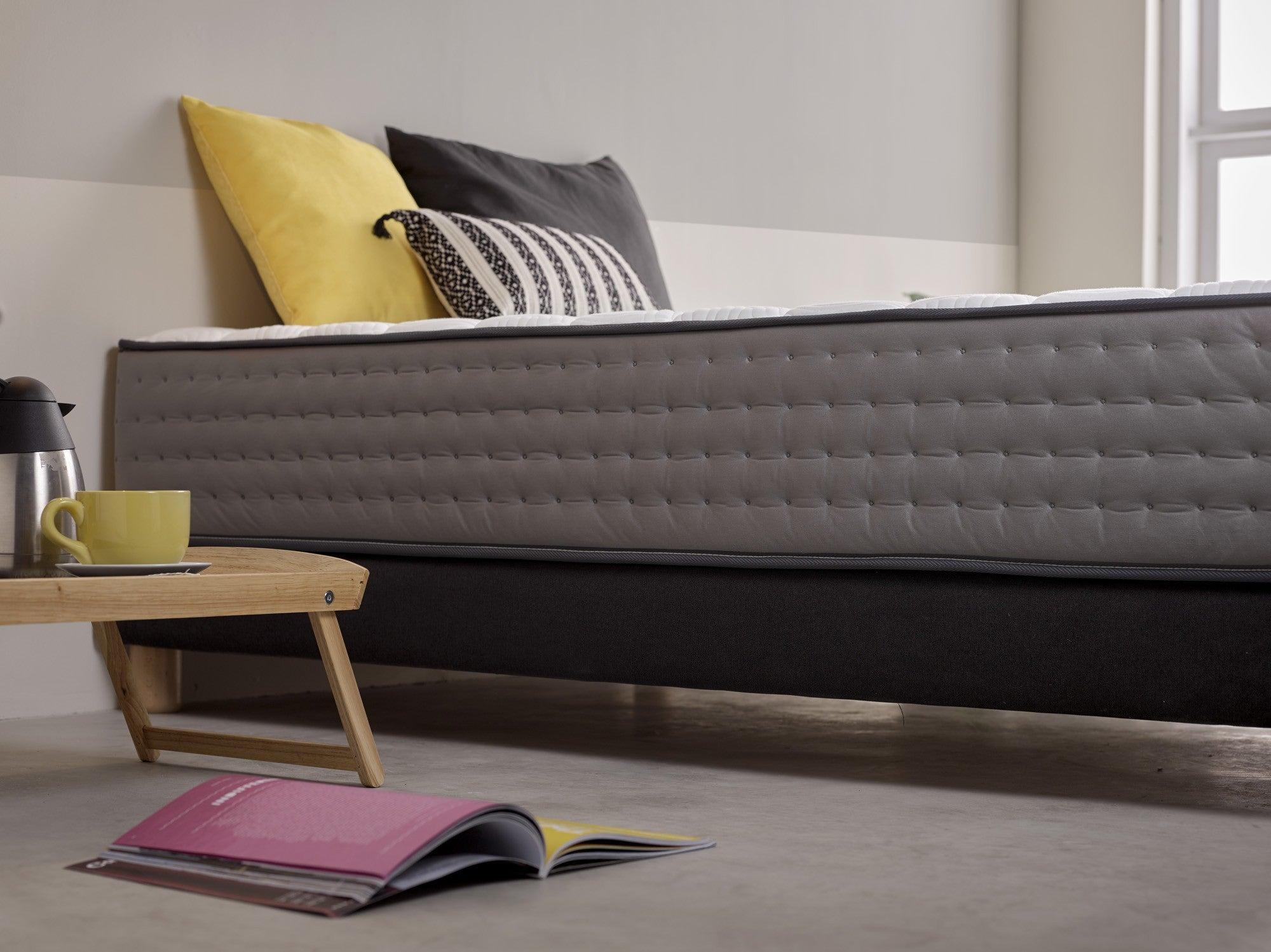 Cama con colchón gris, almohadas y mesa de madera. Se ven un libro y una taza de café.