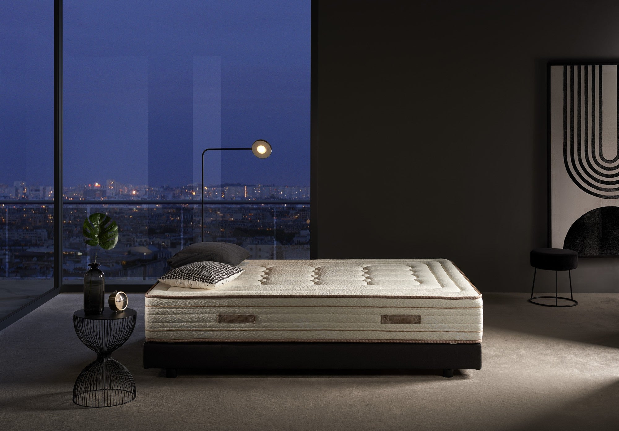 Dormitorio con colchón en estructura de cama, mesa, lámpara y vista de una ciudad de noche.