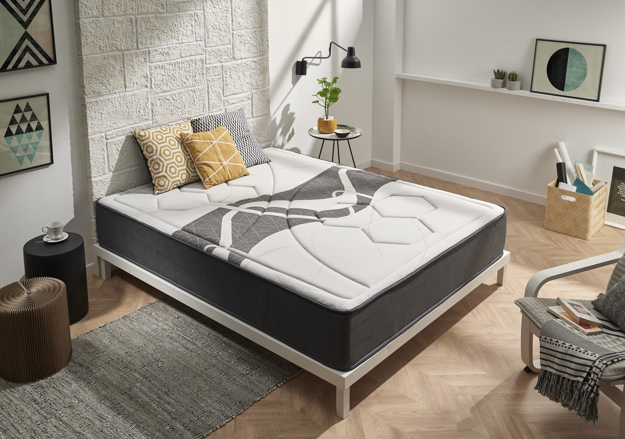 Cama con colchón con borde gris, almohadas y marco blanco. Pared de ladrillo y decoración en el fondo.