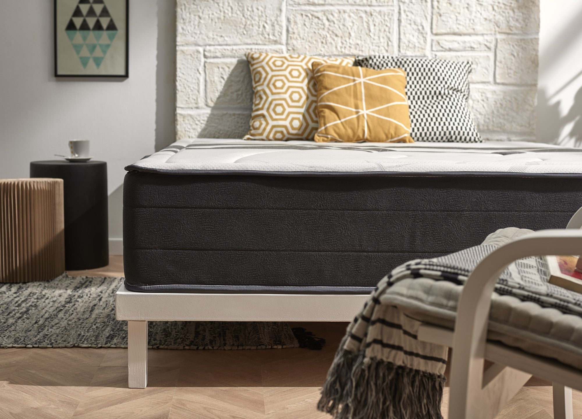 Una cama con colchón blanco, ribete gris y almohadas contra una pared de ladrillos blancos. La cama es central.